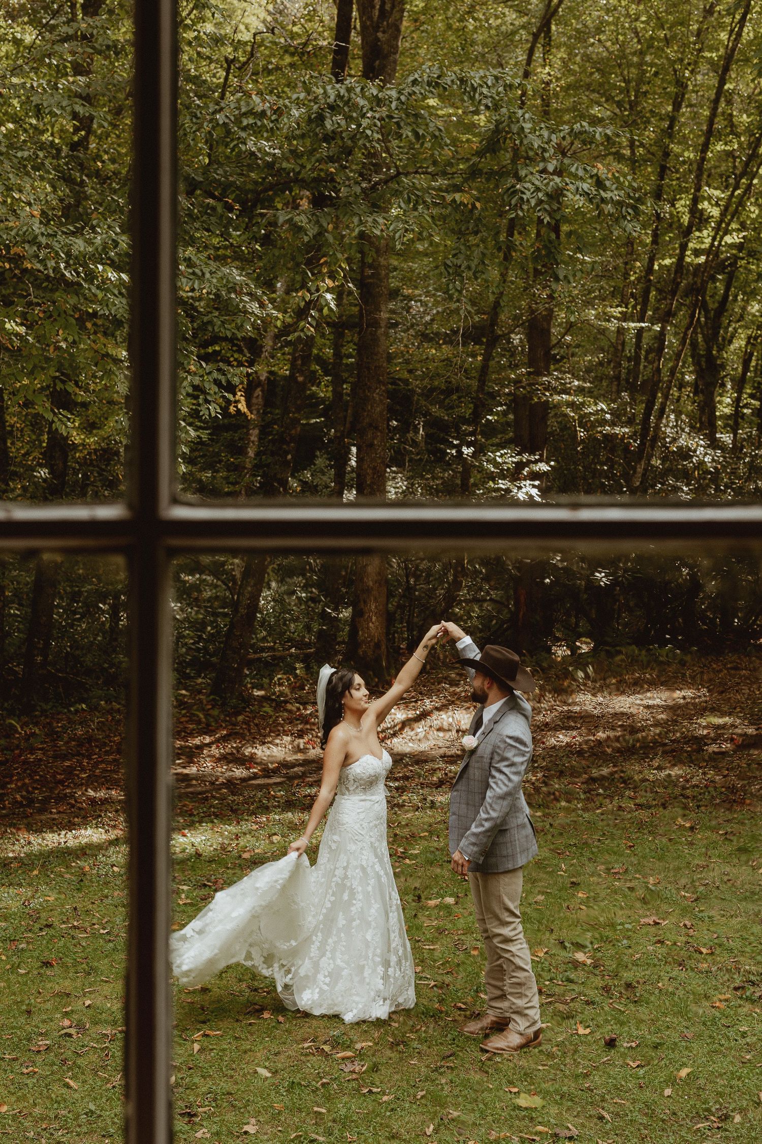 Bride and groom dancing outdoors in a lush forest setting, viewed through a window, bride in white gown.