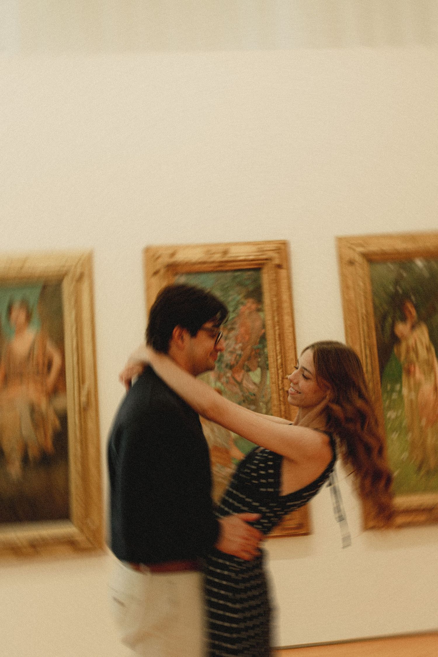 Couple embracing romantically in an art museum gallery with classical framed paintings on the wall behind them.