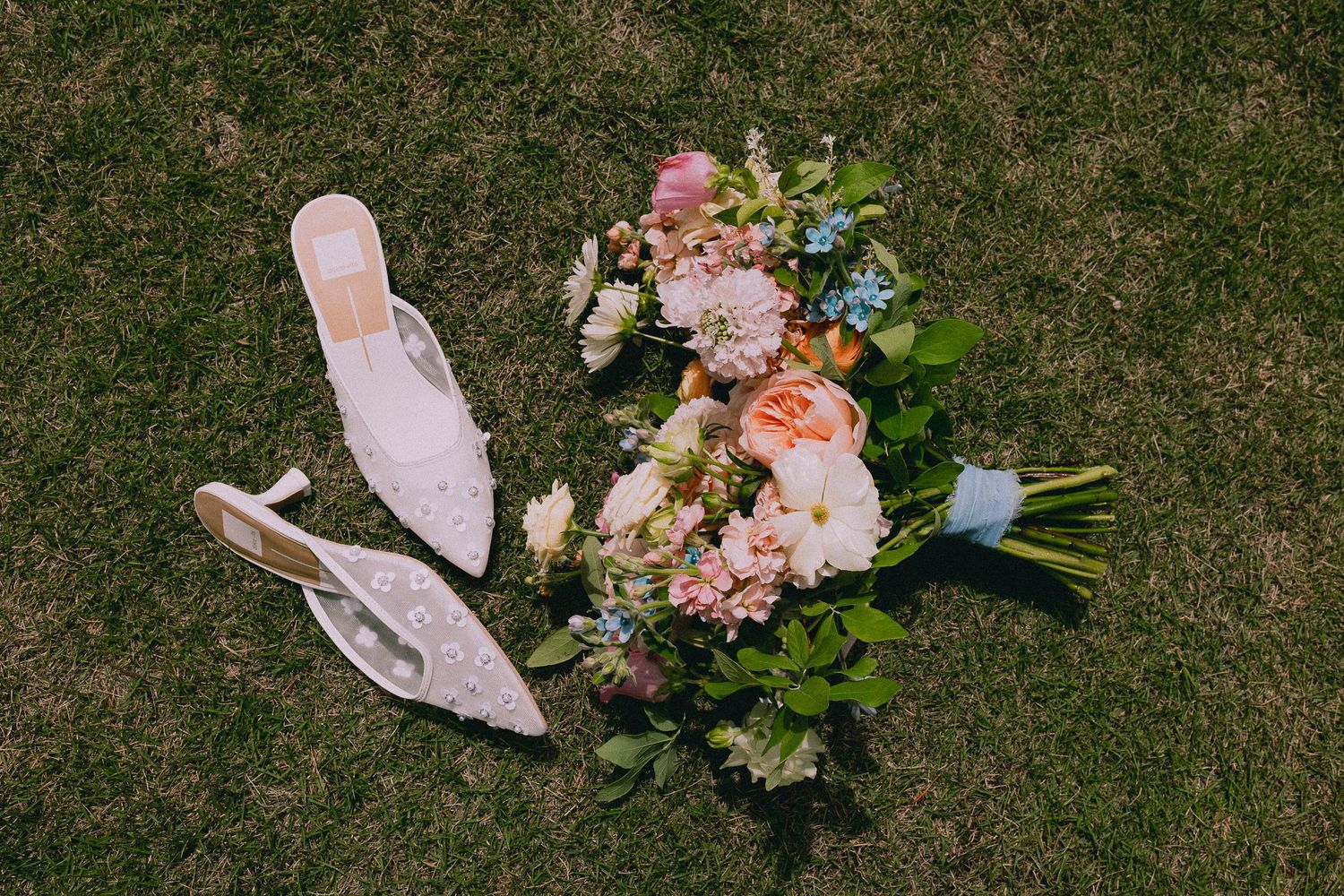 Bridal flat shoes with lace detail beside a colorful wedding bouquet of pink, peach, and blue flowers on green grass.