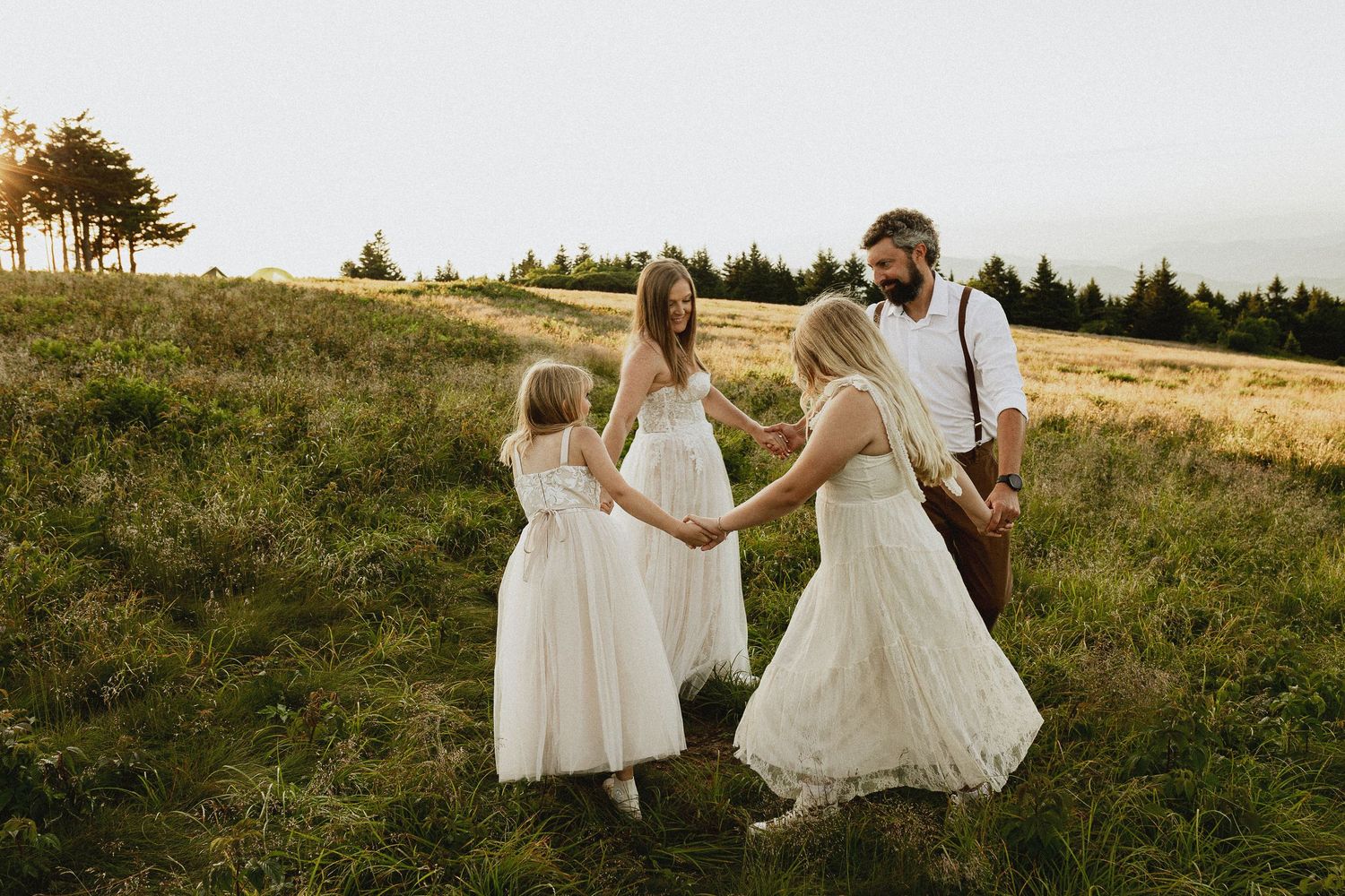 A bearded groom in suspenders holds hands with his bride and two flower girls in white dresses on a green hillside.