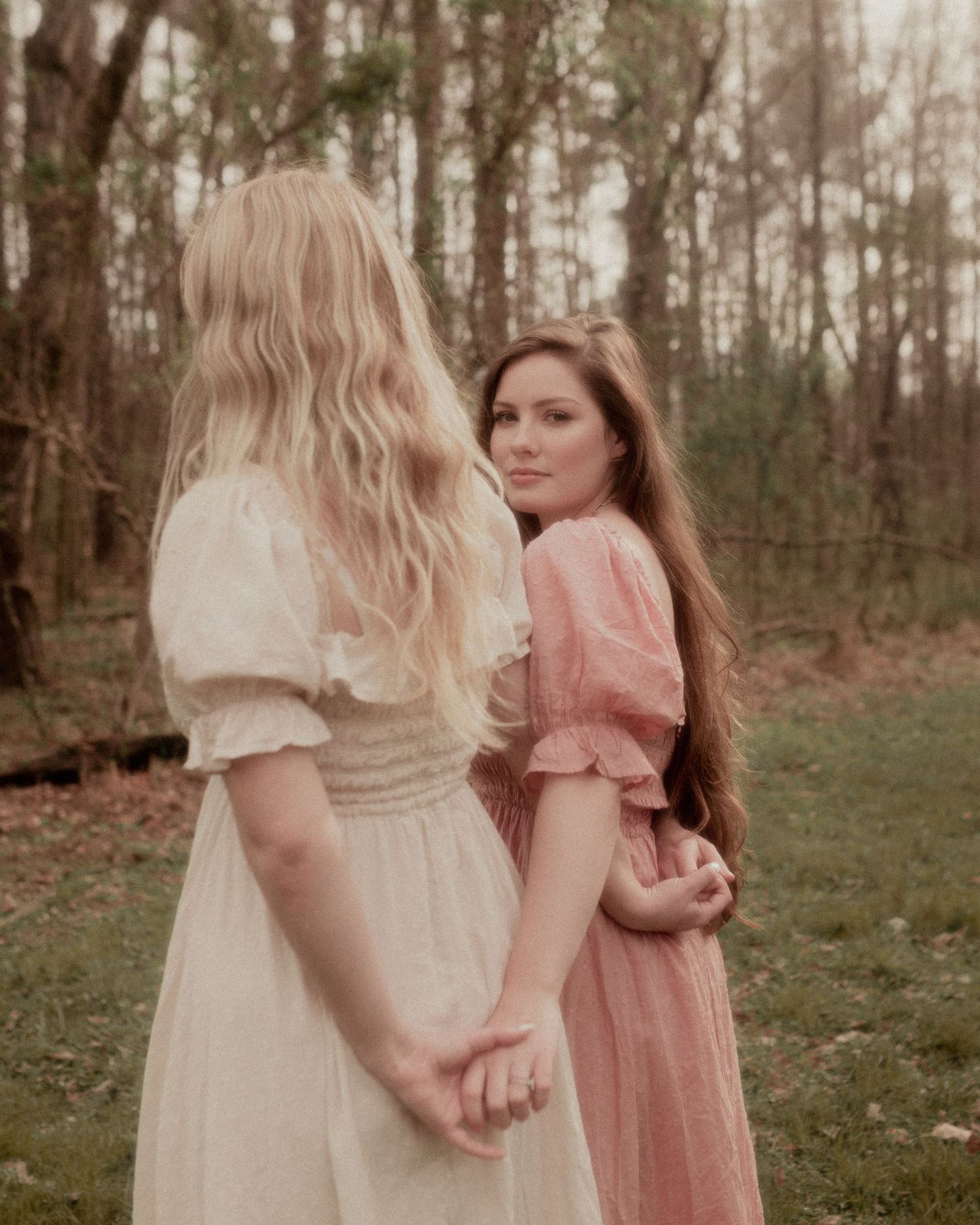 Two people in vintage-style dresses stand together in a dreamy forest setting.