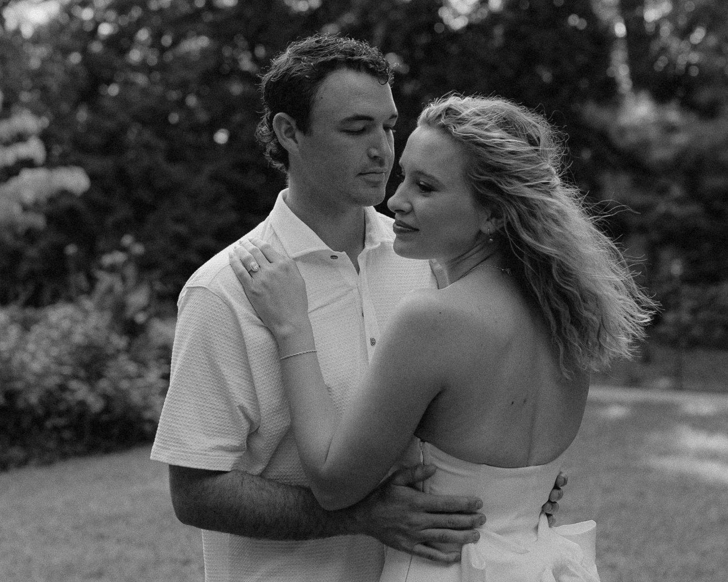 Black and white photo of a romantic couple embracing outdoors, woman in strapless dress, man in white shirt, lush garden background.