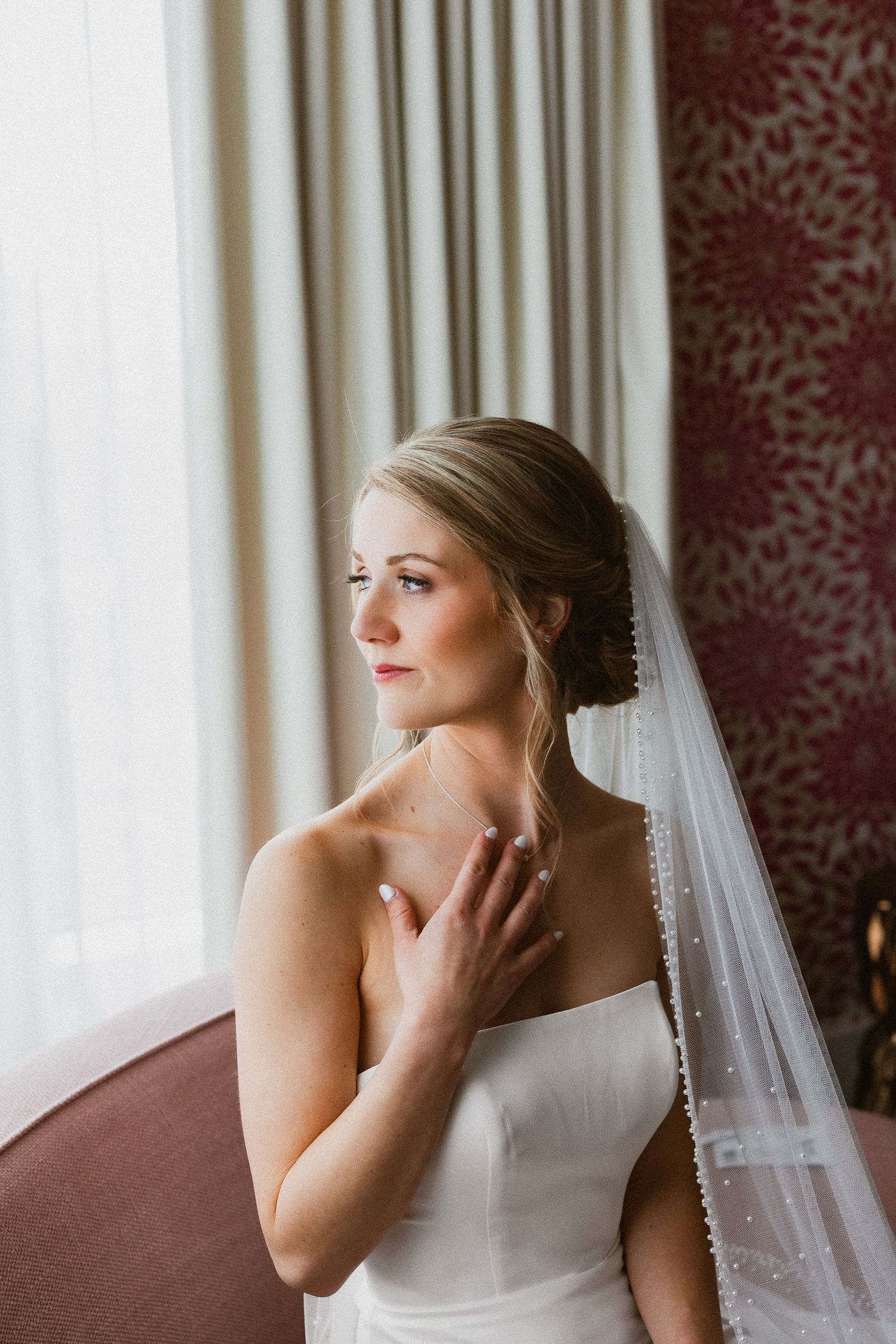 Bride in strapless white gown and pearl-edged veil gazes thoughtfully by a window in an elegant hotel room.