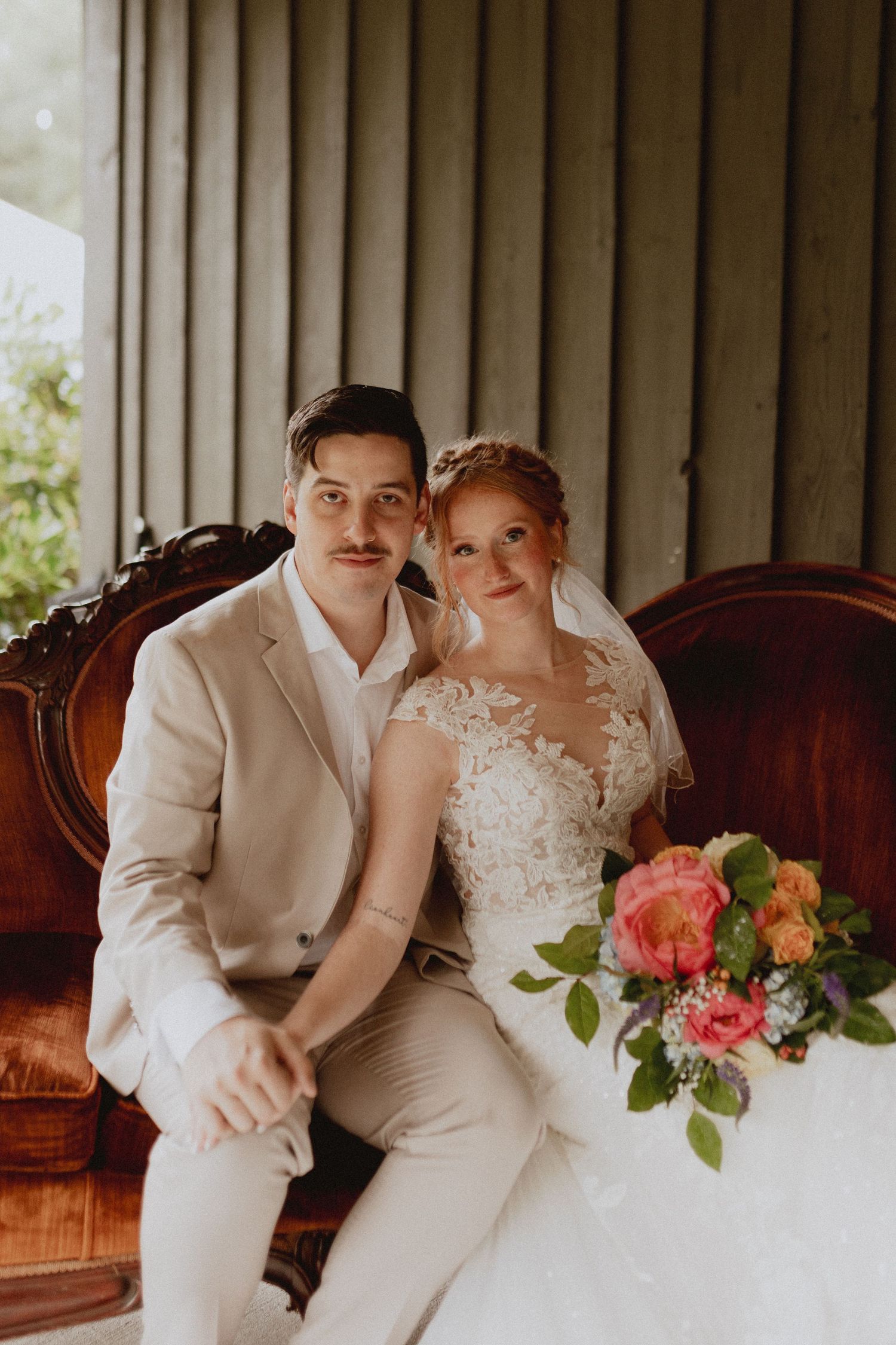 Newlyweds sit close together on vintage sofa with coral and peach floral arrangements.