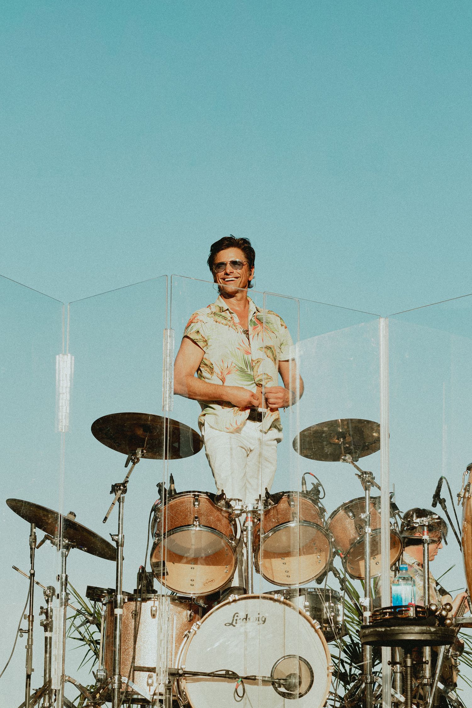 Drummer performs behind extensive drum kit setup against bright blue sky background.