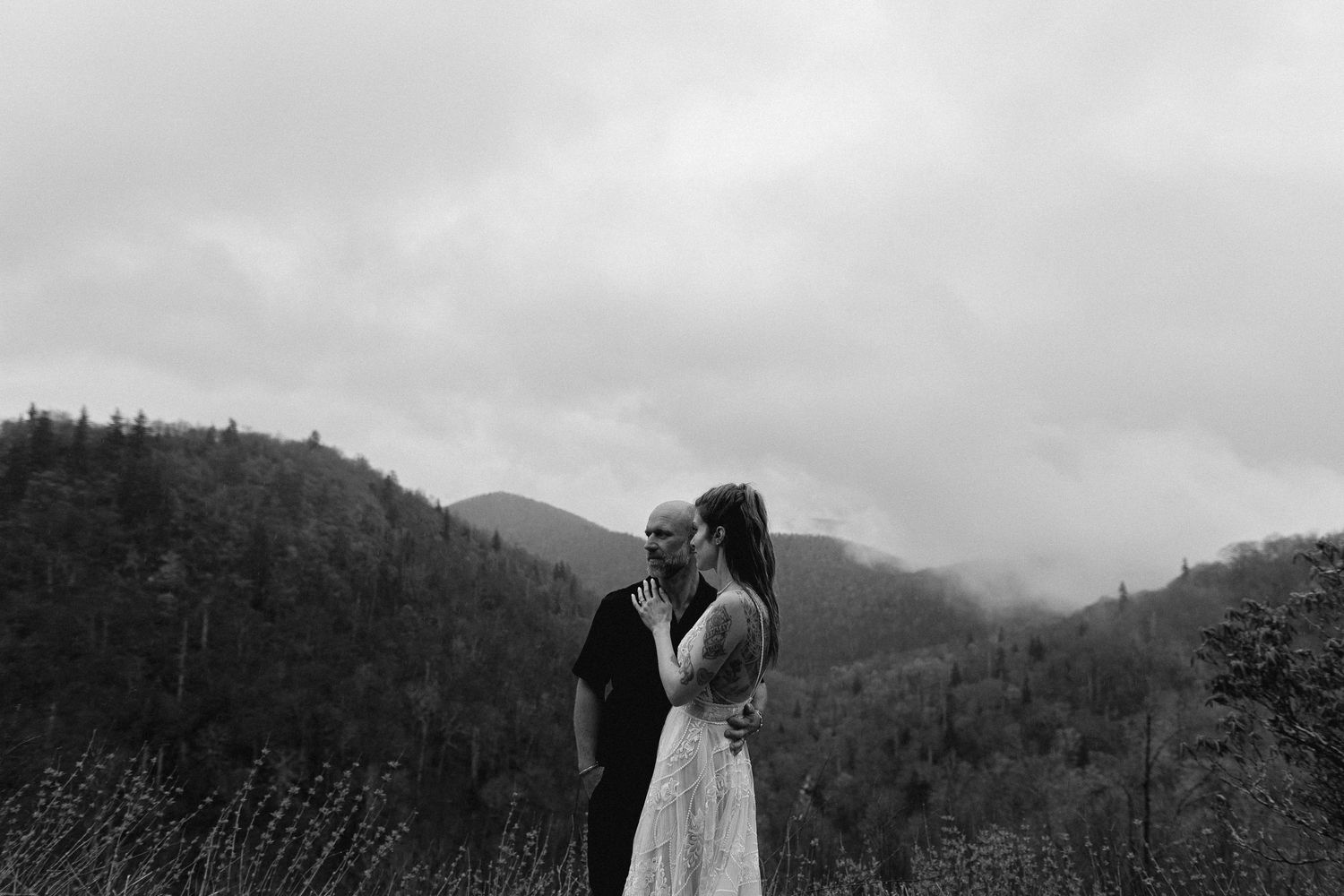 A black and white photo captures a wedding couple against a mountainous landscape on an overcast day.