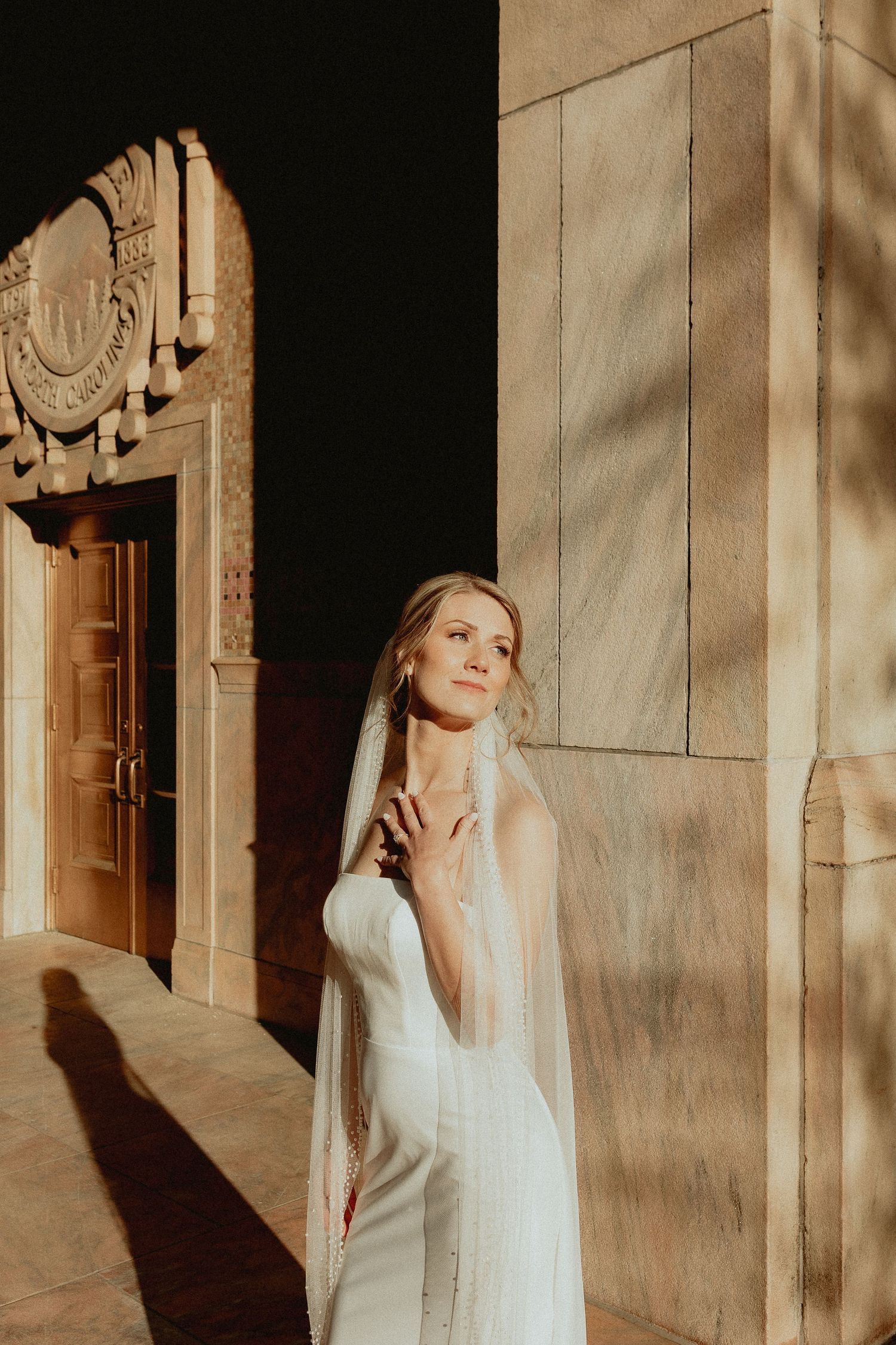 Bride in white gown and veil poses gracefully outside ornate stone building in golden sunlight.