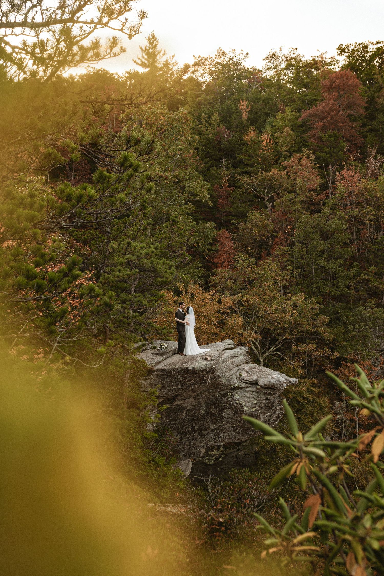 Distant wedding couple stands on rocky outcrop surrounded by autumn foliage and golden light.