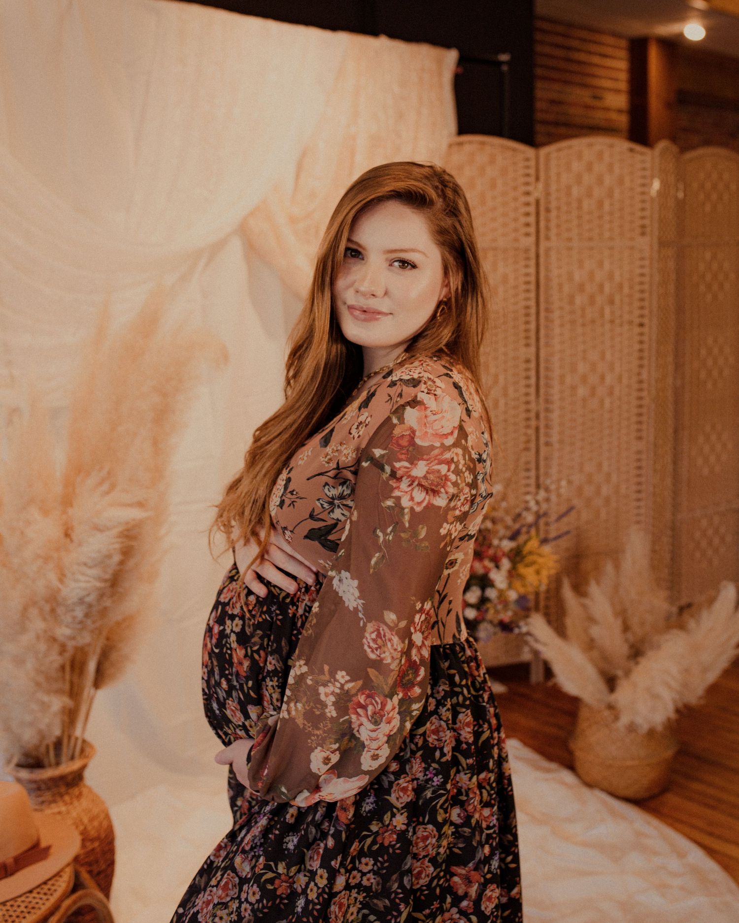 Woman in floral dress poses against warm-toned interior backdrop.