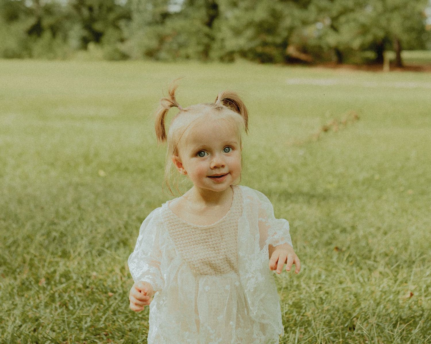 A toddler in white dress smiles while playing outdoors in a park.
