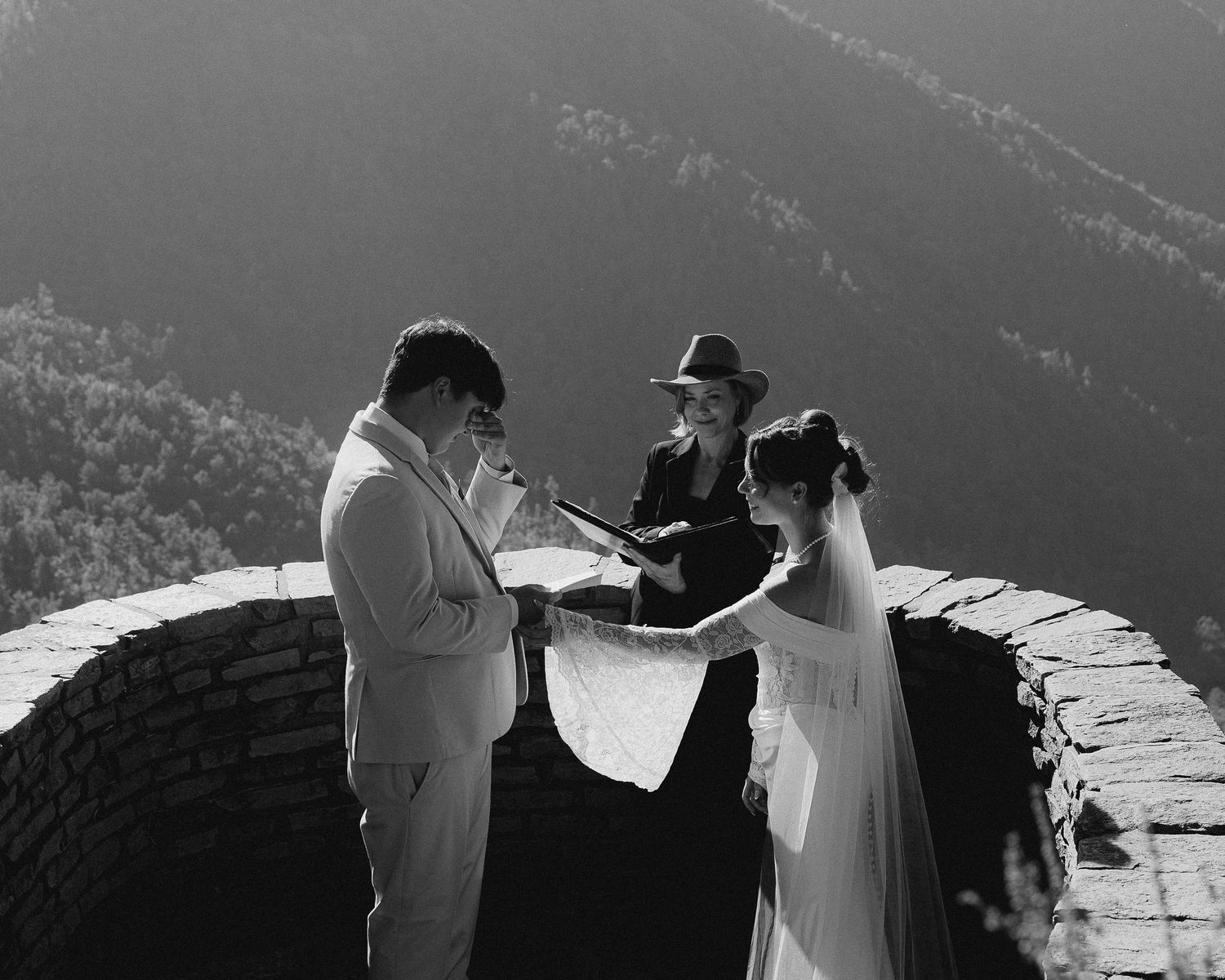 Black and white photo of a couple exchanging vows with an officiant on a stone overlook with mountain views.
