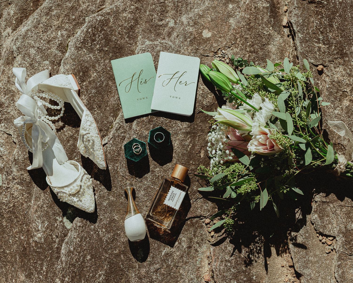 Flat lay of wedding details including lace heels, His and Hers vows, rings, perfume, and floral bouquet on stone.