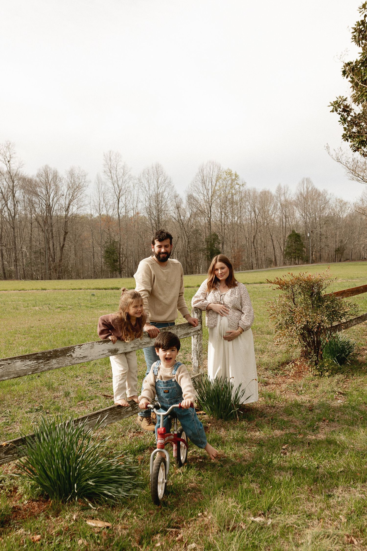 Pregnant woman in white dress with family outdoors, man, two children, one on bicycle, near wooden fence and green field.
