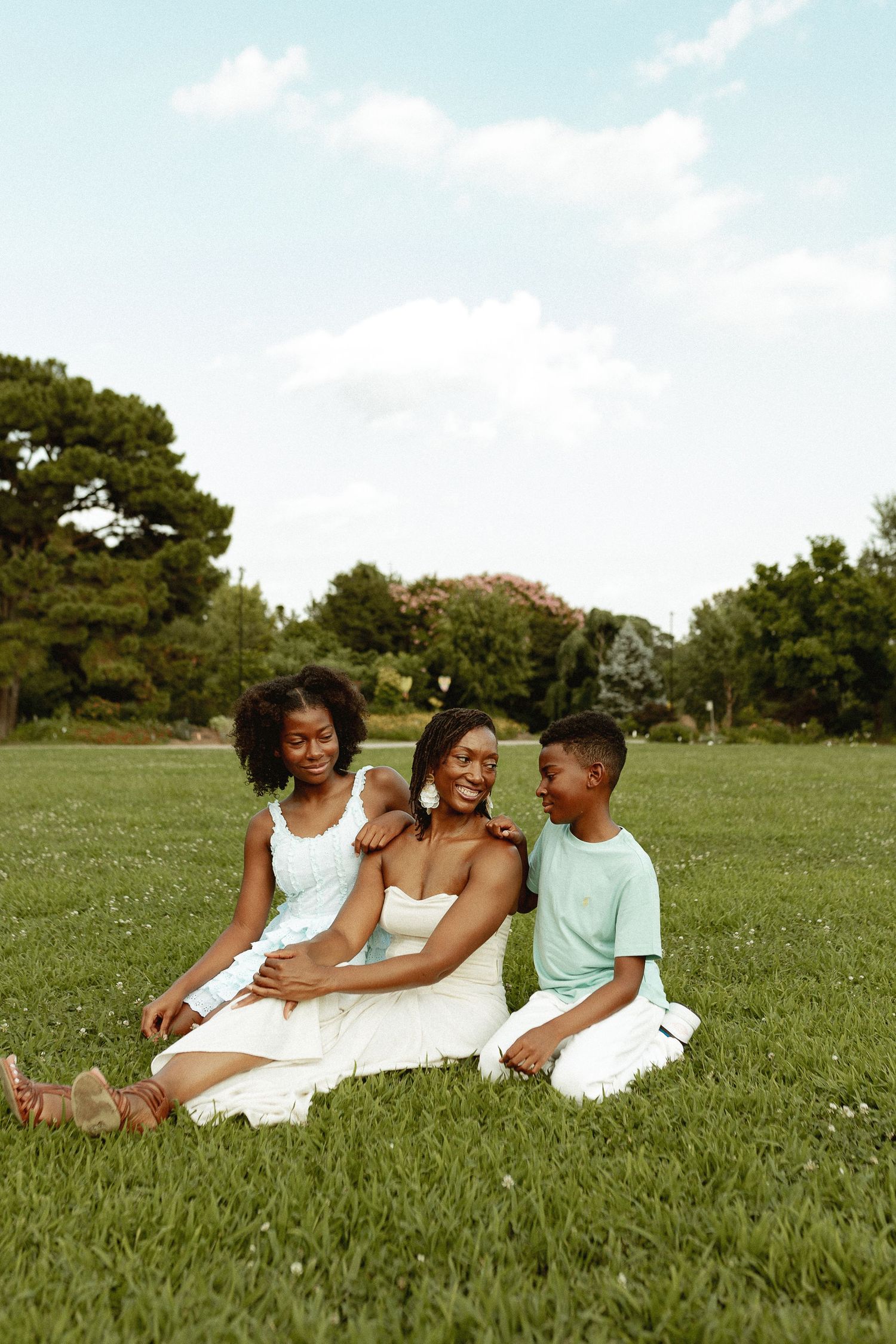 Three people sitting together on grass in a sunny park with trees in the background.