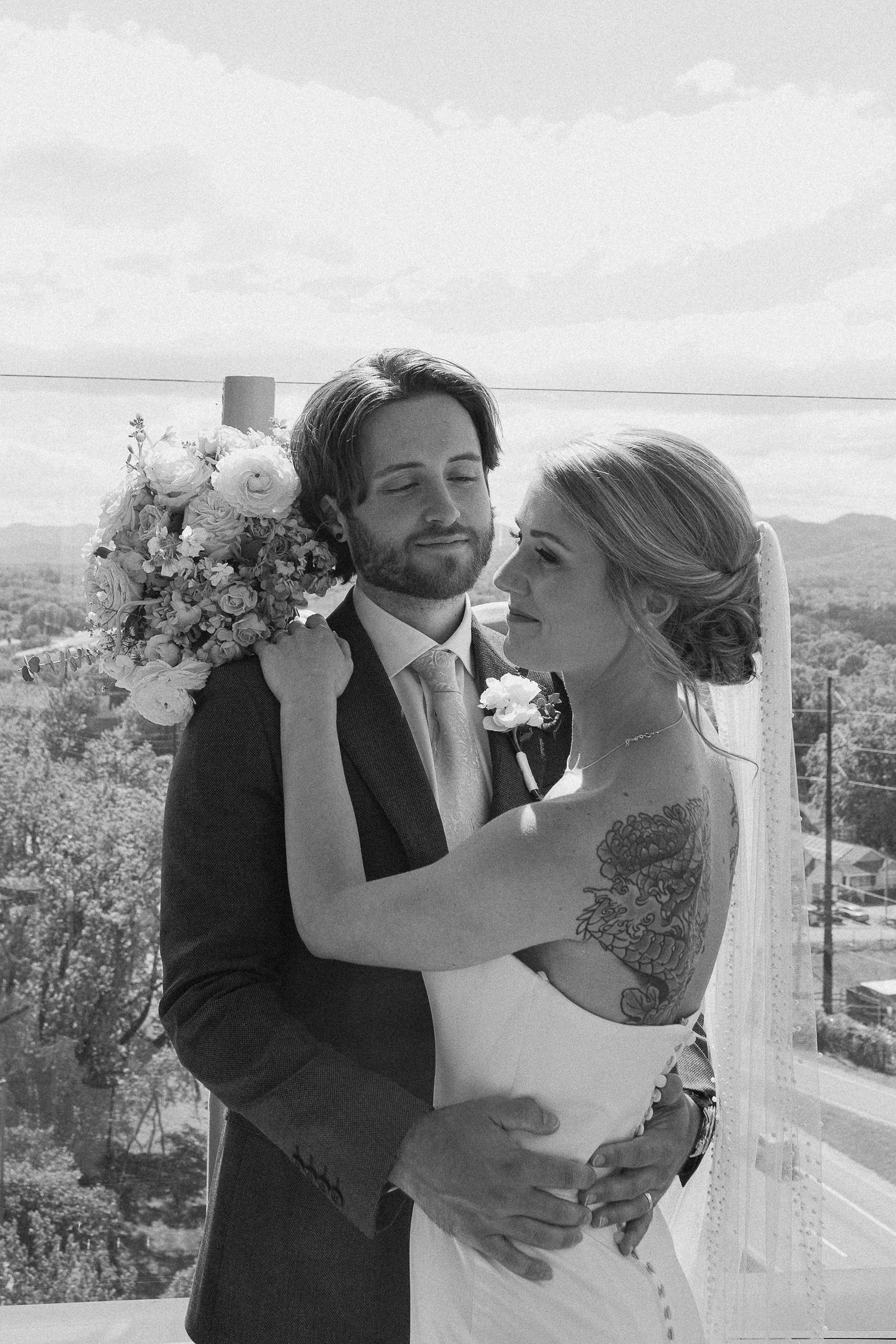 Black and white wedding photo of bride and groom embracing on balcony with floral bouquet and scenic view.