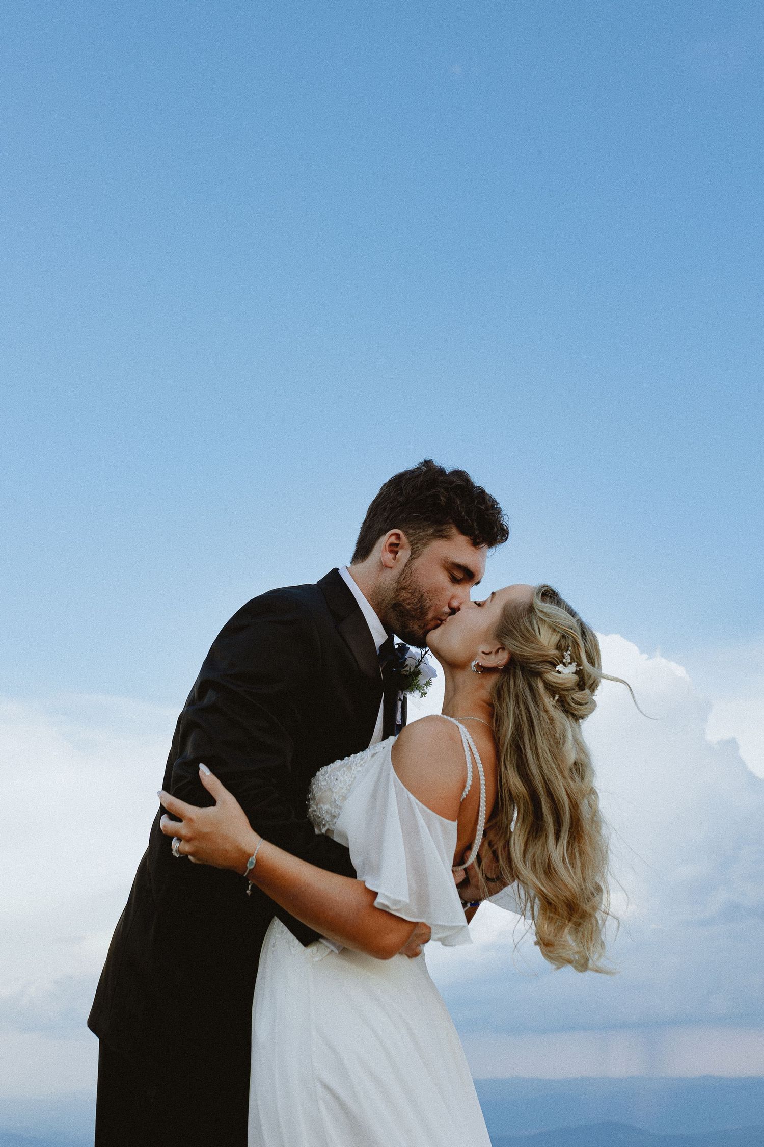 Bride and groom share a romantic kiss outdoors in wedding attire against a blue sky backdrop.