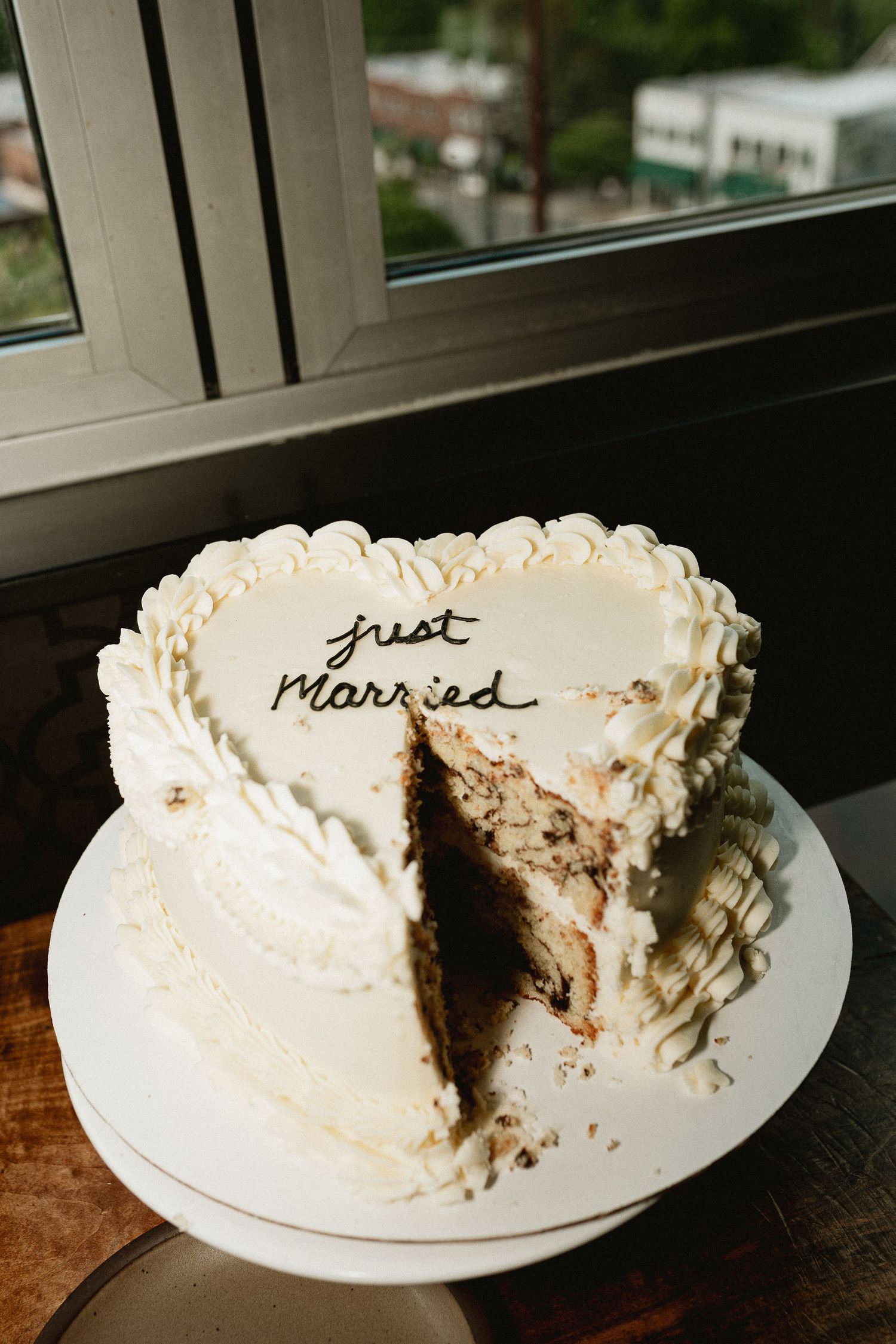 Heart-shaped white wedding cake with Just Married inscription, partially sliced, displayed on white cake stand near window.