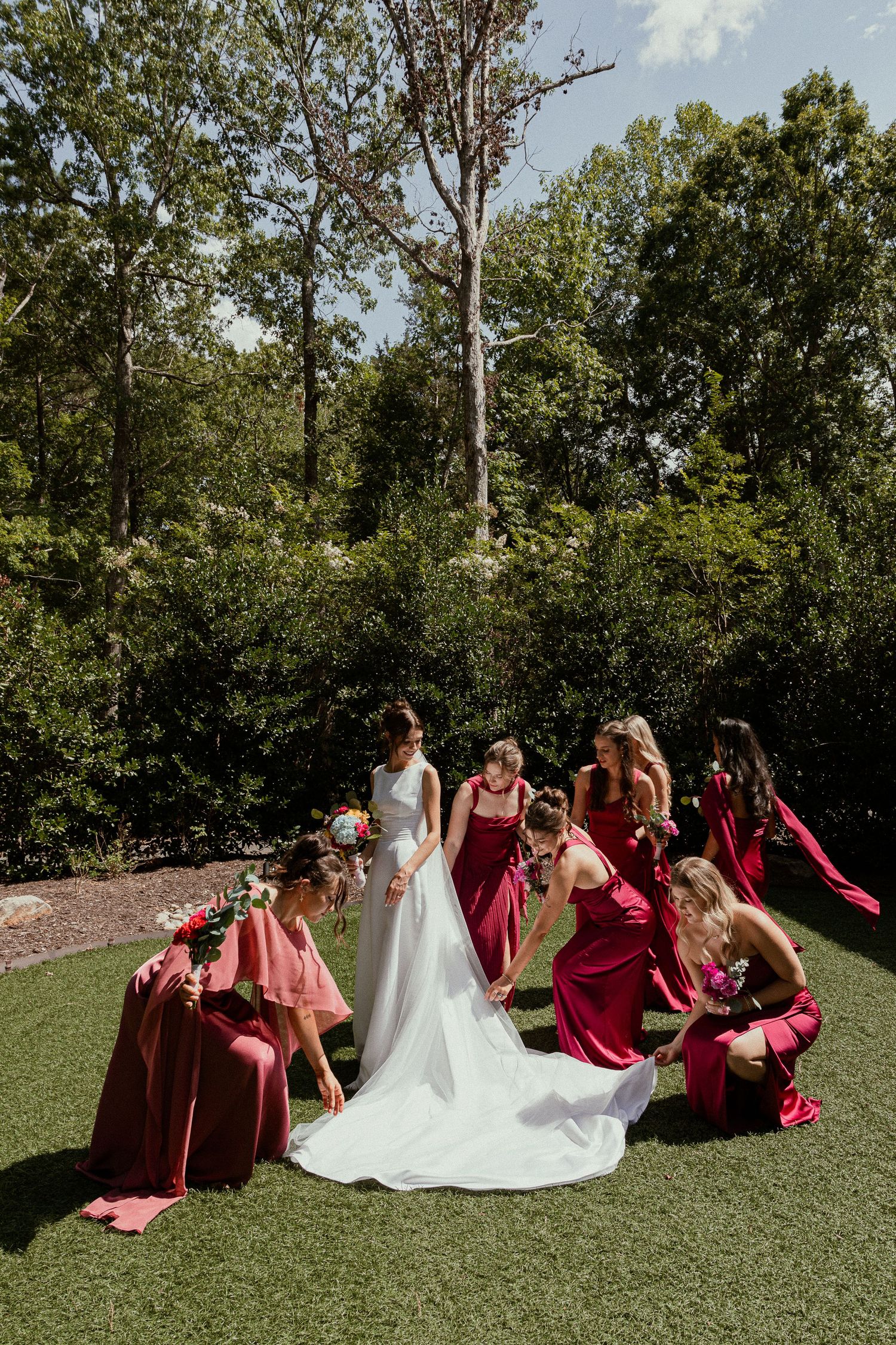Bride in white gown surrounded by bridesmaids in burgundy dresses arranging her train on green lawn with trees behind.