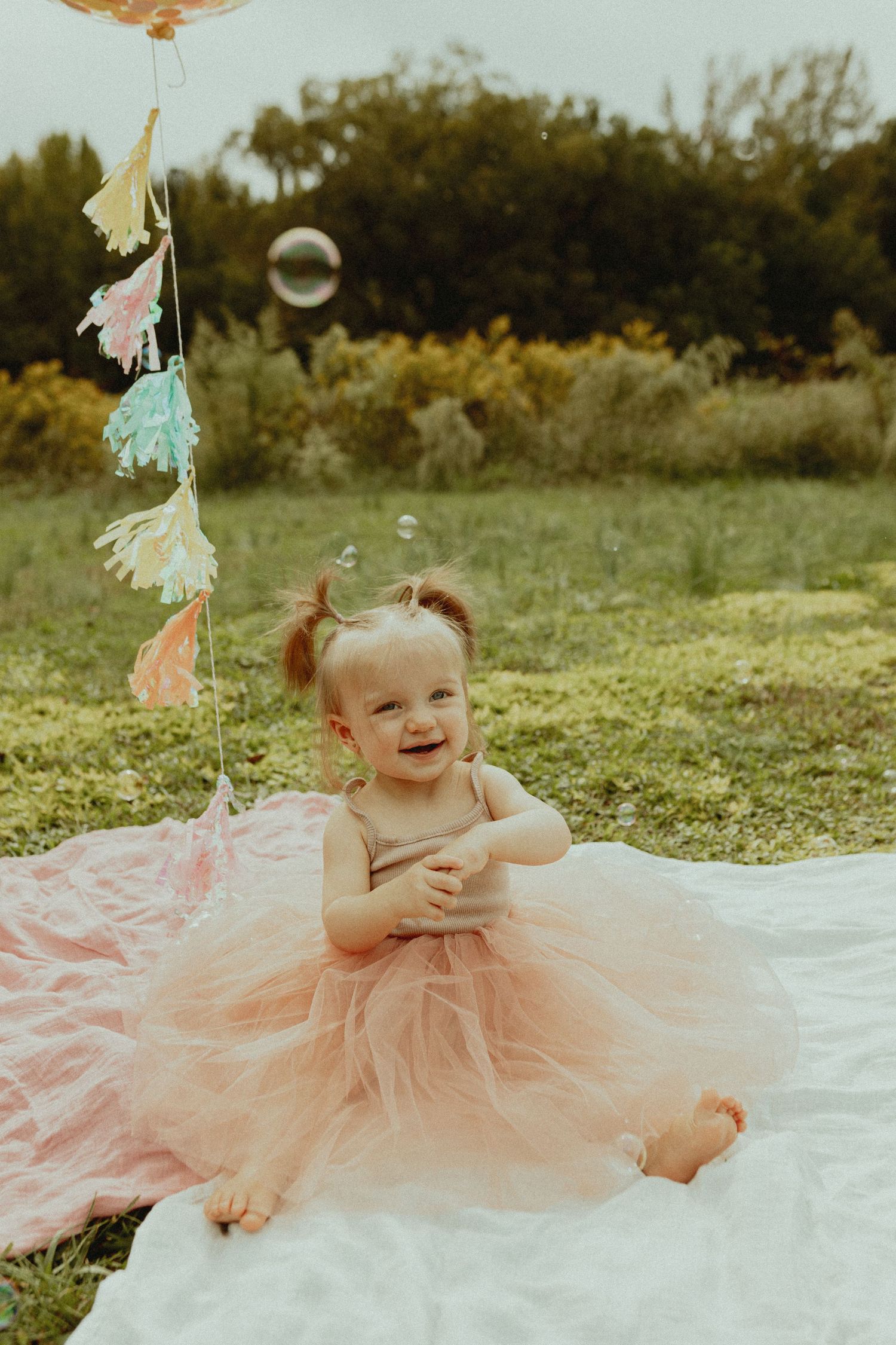 A happy baby in pink tulle dress plays on a white blanket surrounded by decorative tassels.