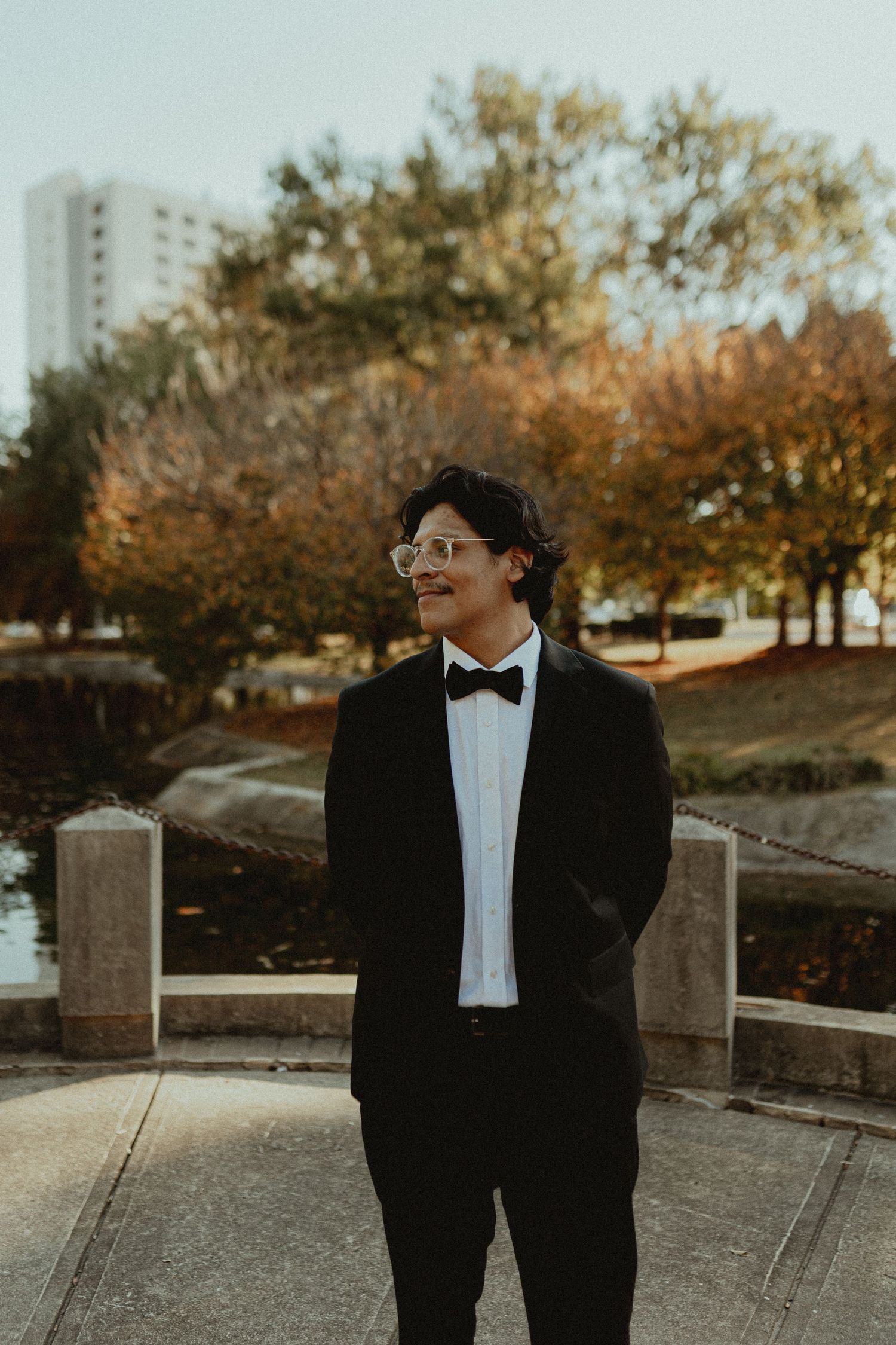 Person in formal black tuxedo poses thoughtfully in an outdoor autumn setting.
