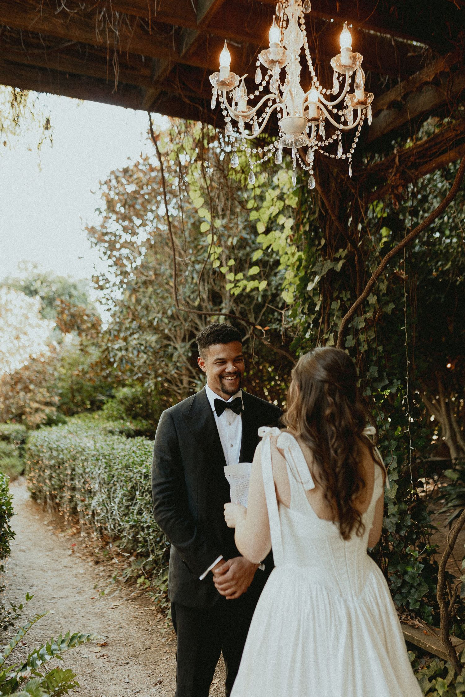 Elegant wedding couple stands beneath crystal chandelier on garden path surrounded by lush greenery.