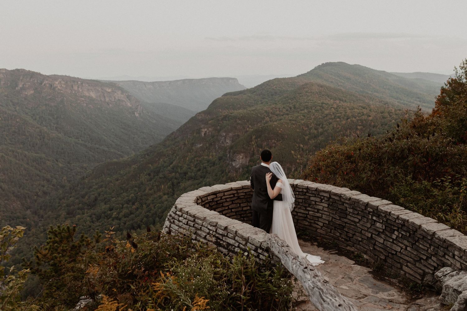 Newlyweds share an intimate moment on stone wall overlooking mountain vista.