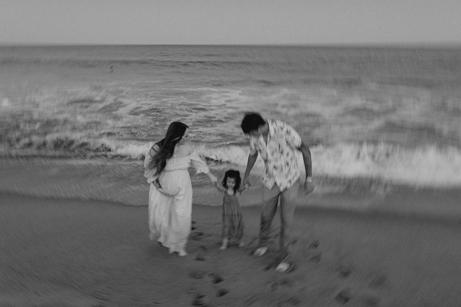 Black and white beach scene with silhouettes walking along the shoreline at sunset.
