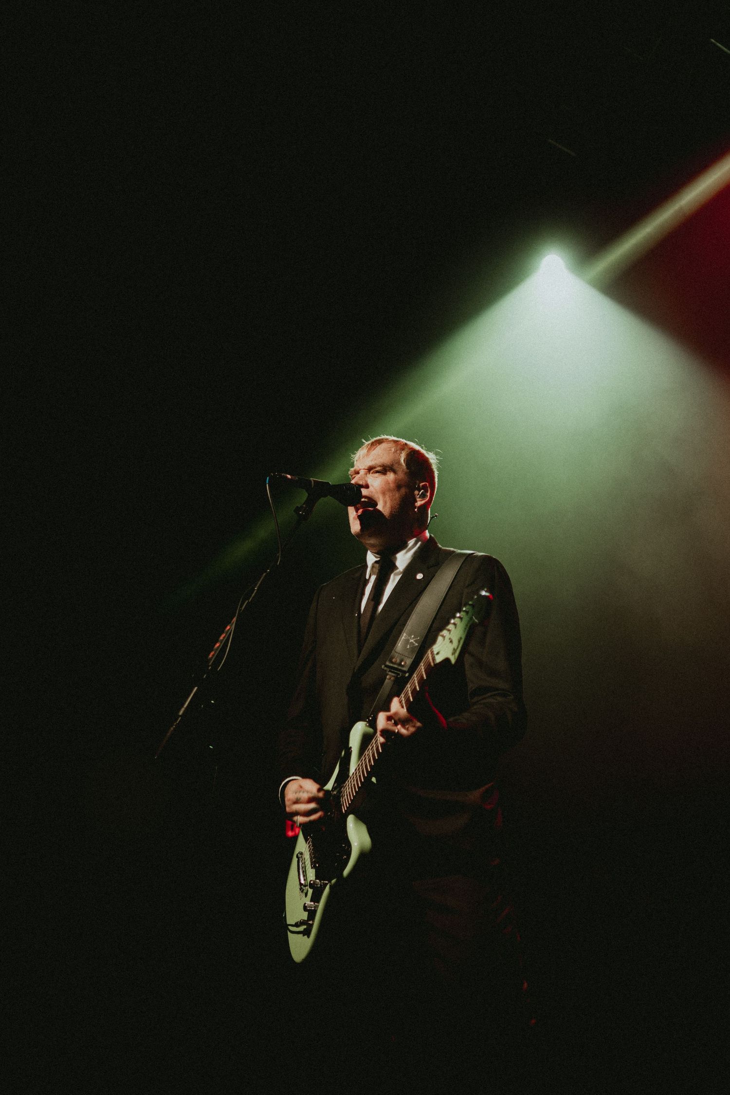 Musician in suit performs with guitar under dramatic spotlight on dark stage.