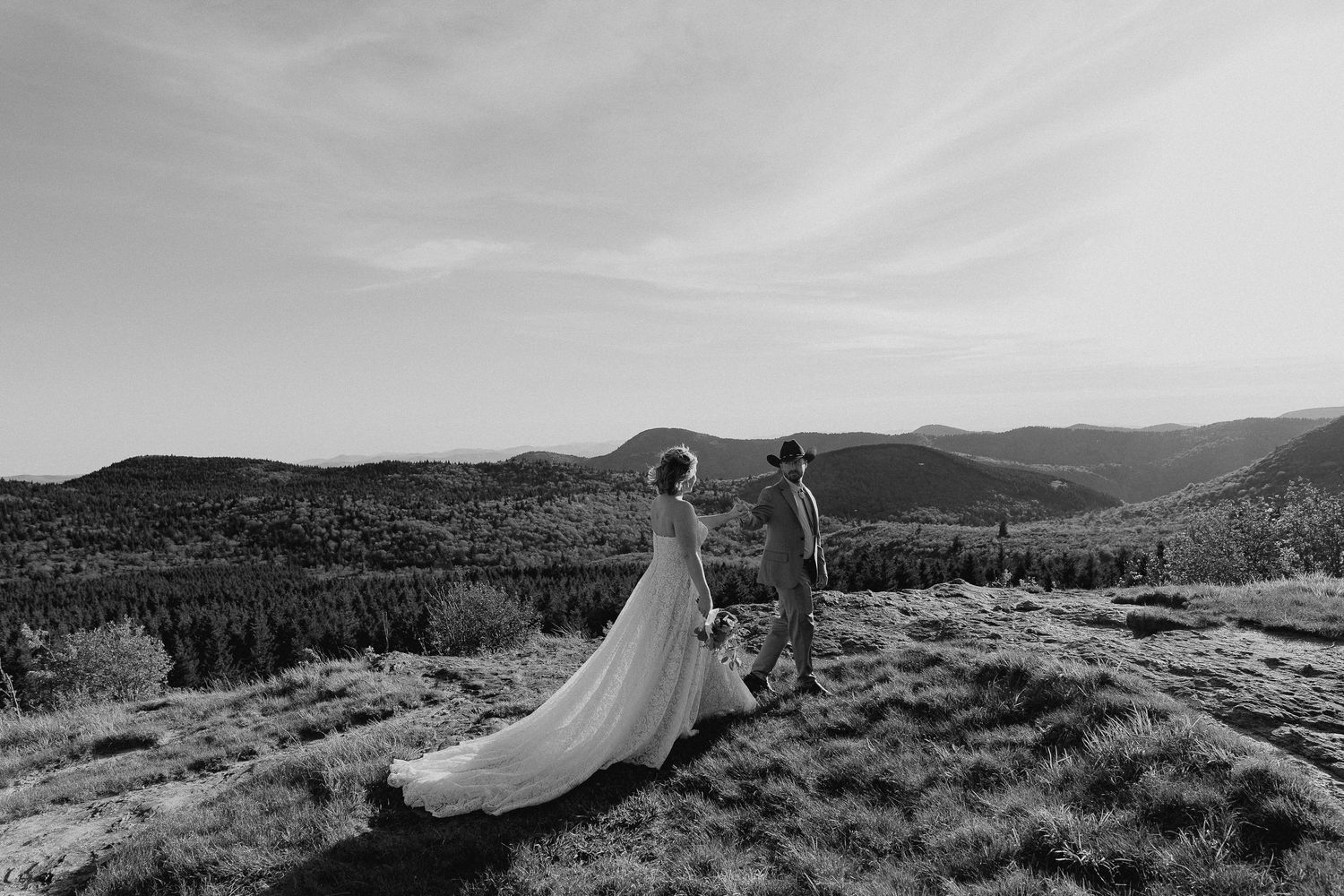 Black and white photo of a bride and groom holding hands on a mountain summit with sweeping valley views.