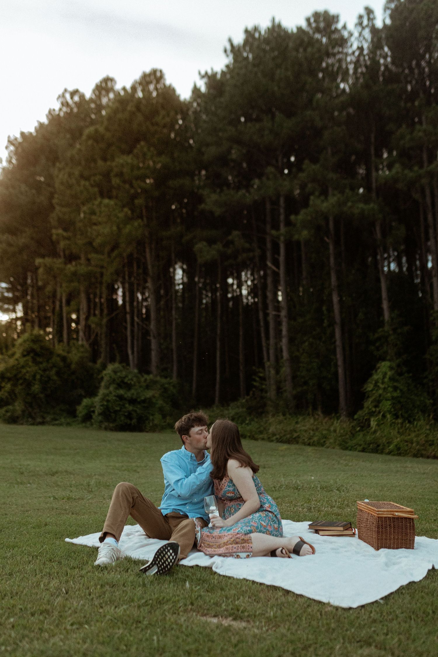 A couple enjoys a romantic picnic on a blanket in a grassy field near pine trees.
