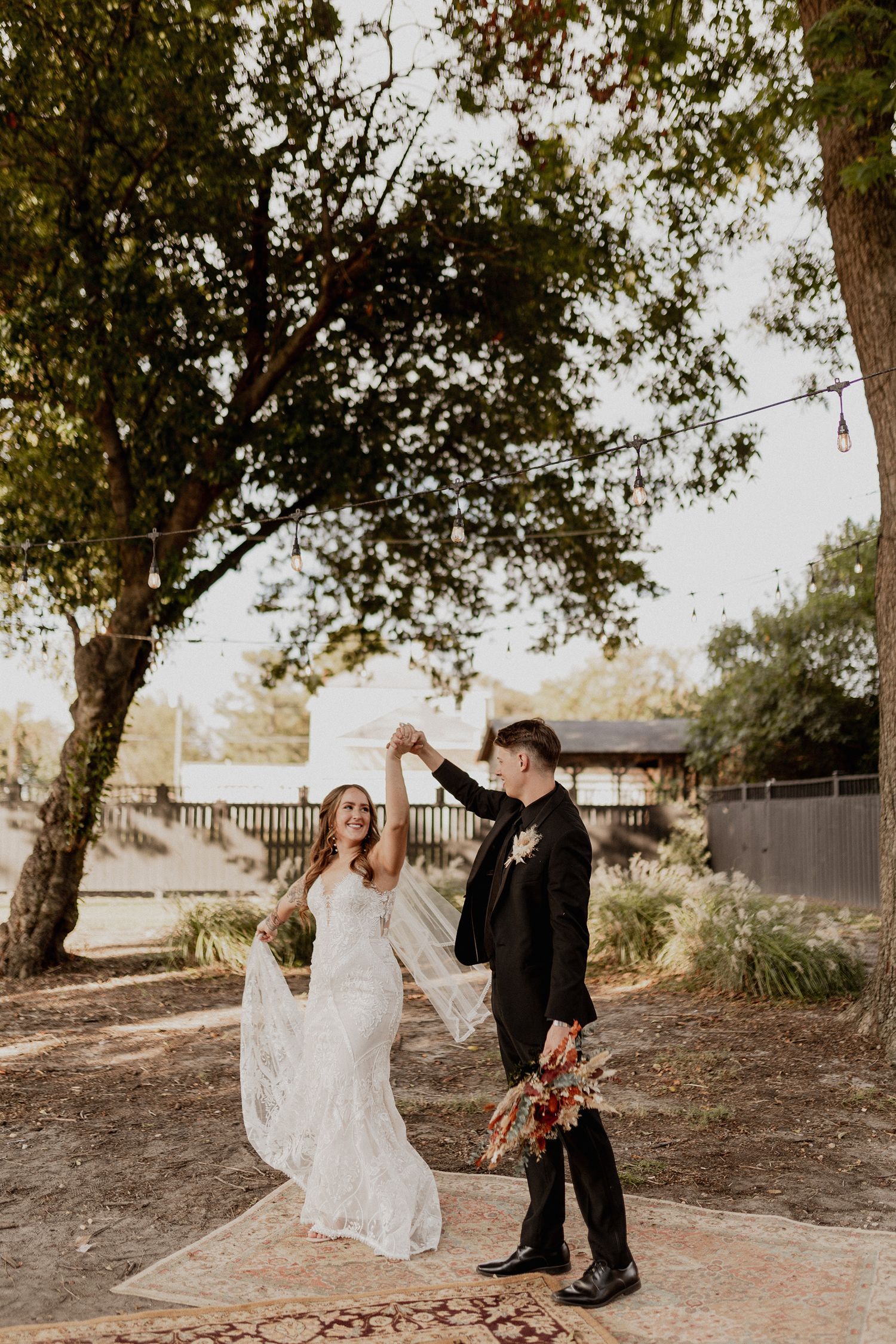 A couple in wedding attire shares a playful moment under trees in natural lighting.