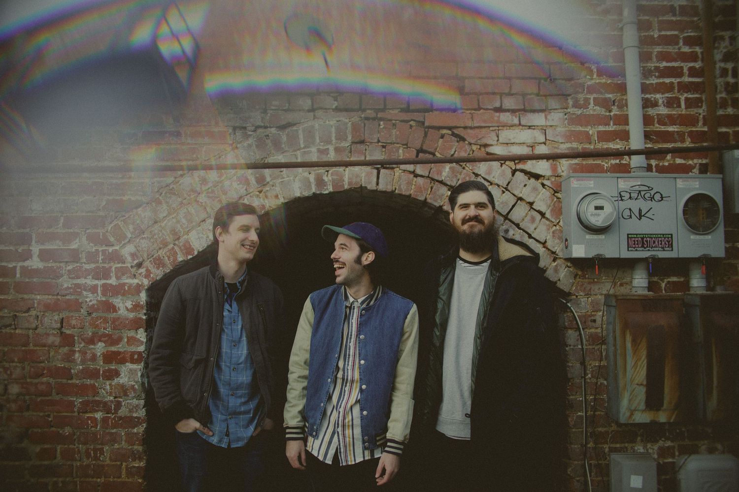 Three musicians stand in front of a brick archway in casual attire.