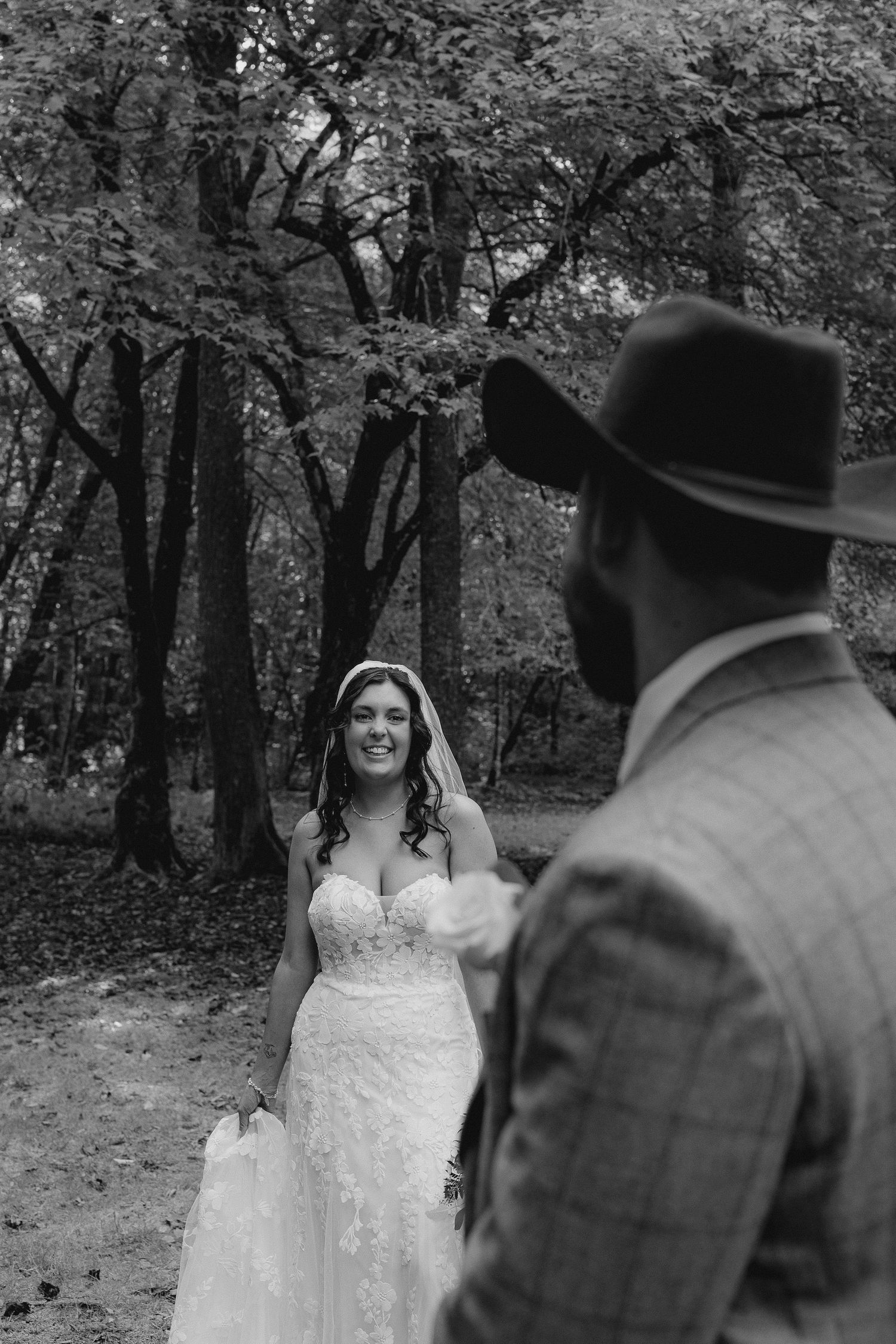 Black and white photo of a bride in lace gown smiling at groom wearing cowboy hat during outdoor forest first look.