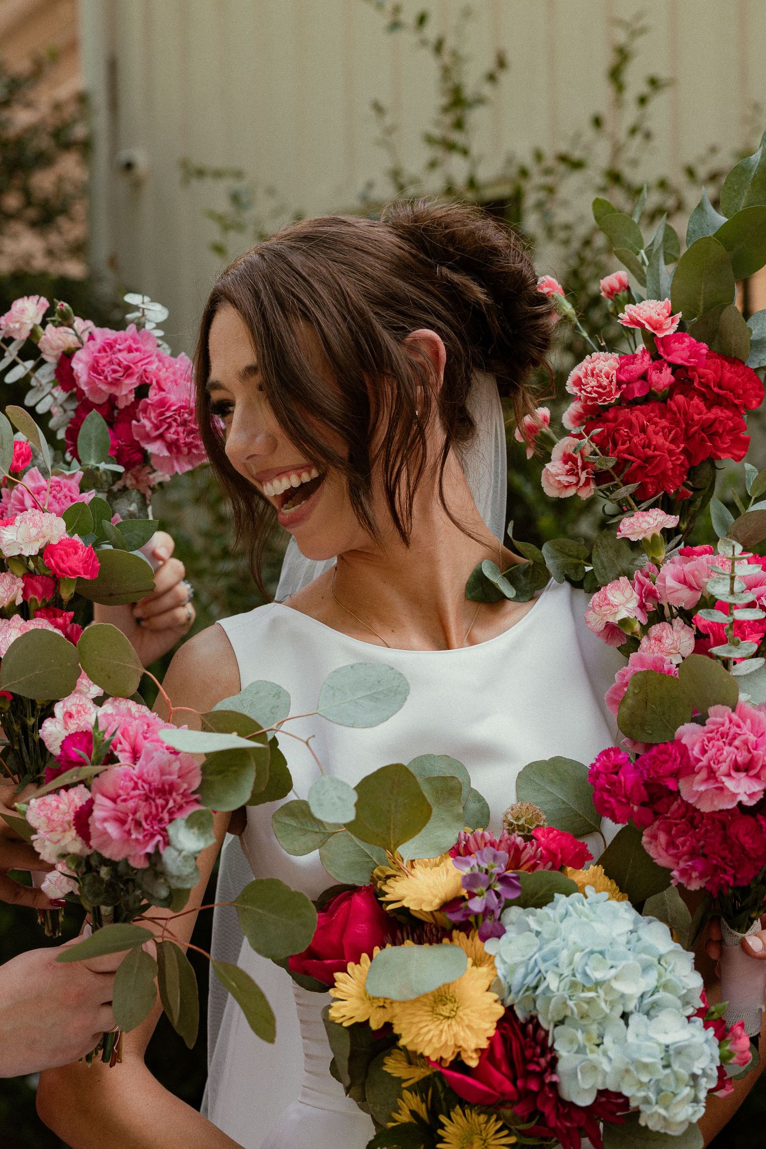 Smiling bride in white dress surrounded by vibrant pink, red, and yellow flower bouquets with eucalyptus greenery.