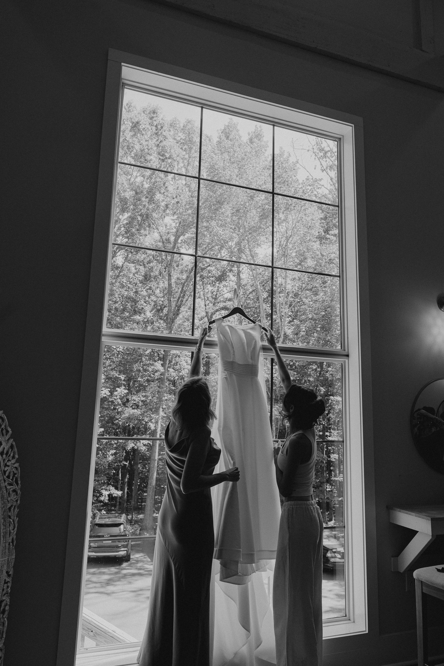 Two bridesmaids hang a white wedding dress on a large window in a black and white photo.