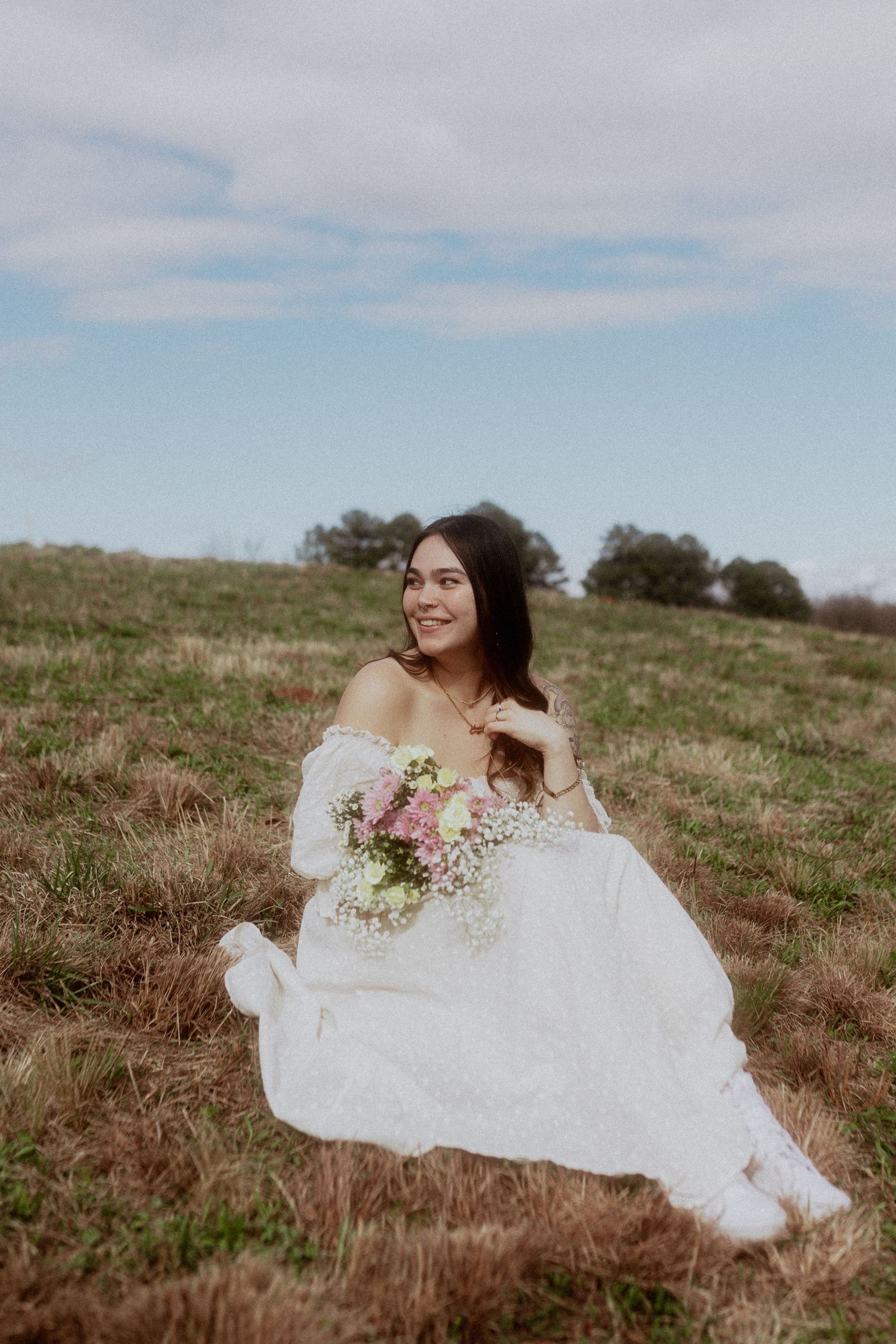 Person in white wedding dress lying in field with flowers under cloudy sky.