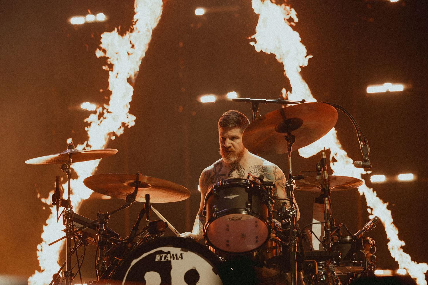Drummer performs behind drum kit with fire pyrotechnics shooting up in background.