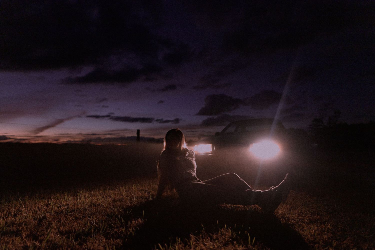 Silhouette of person sitting in field at night with glowing lights in background.