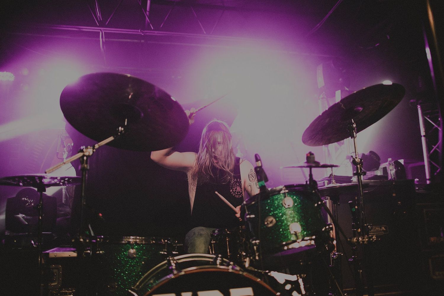 Purple stage lights illuminate a concert venue with drum kit silhouettes visible.