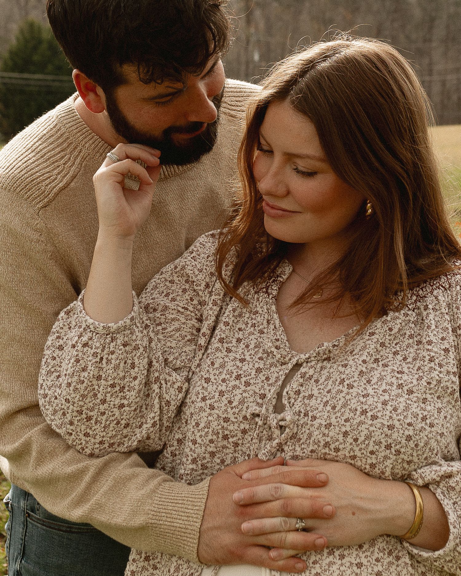 A couple shares a tender moment outdoors, the woman in a floral dress cradling her baby bump as her bearded partner looks on lovingly.