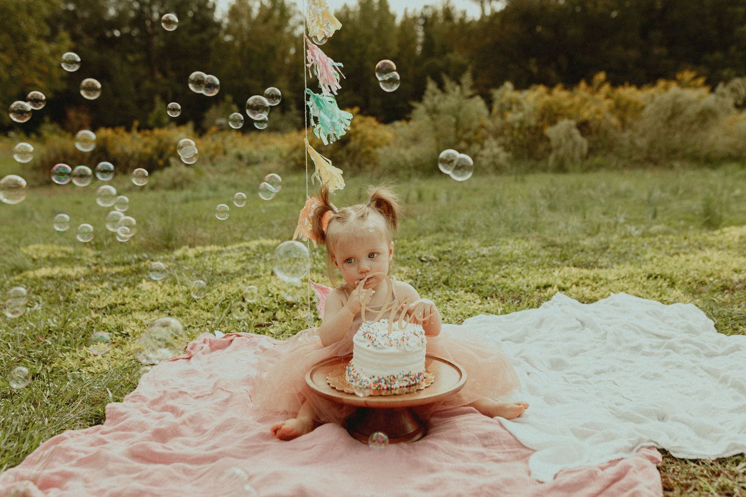 A baby in pink dress plays with bubbles on a white blanket outdoors.
