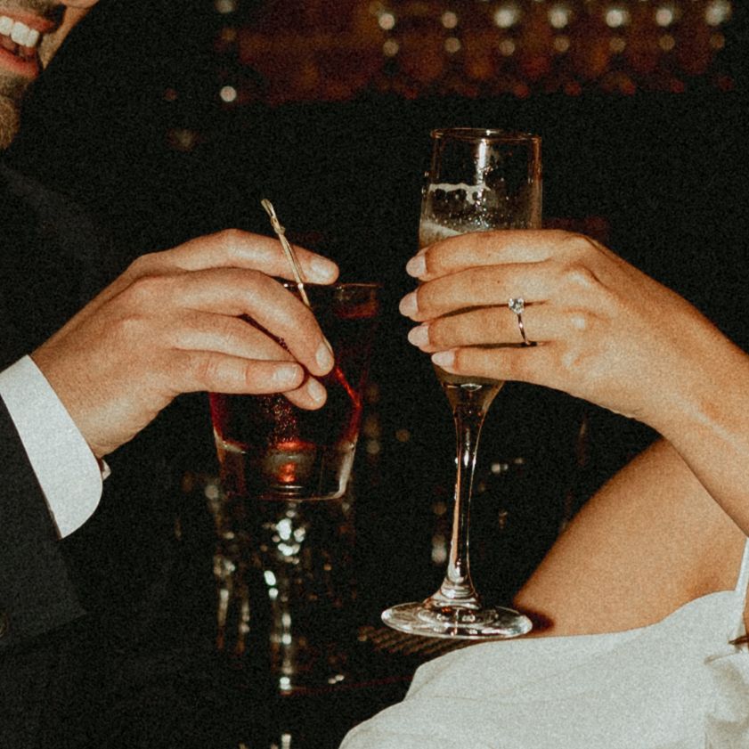 Hands holding wine glasses clinking together in a celebratory toast at a dimly lit venue.