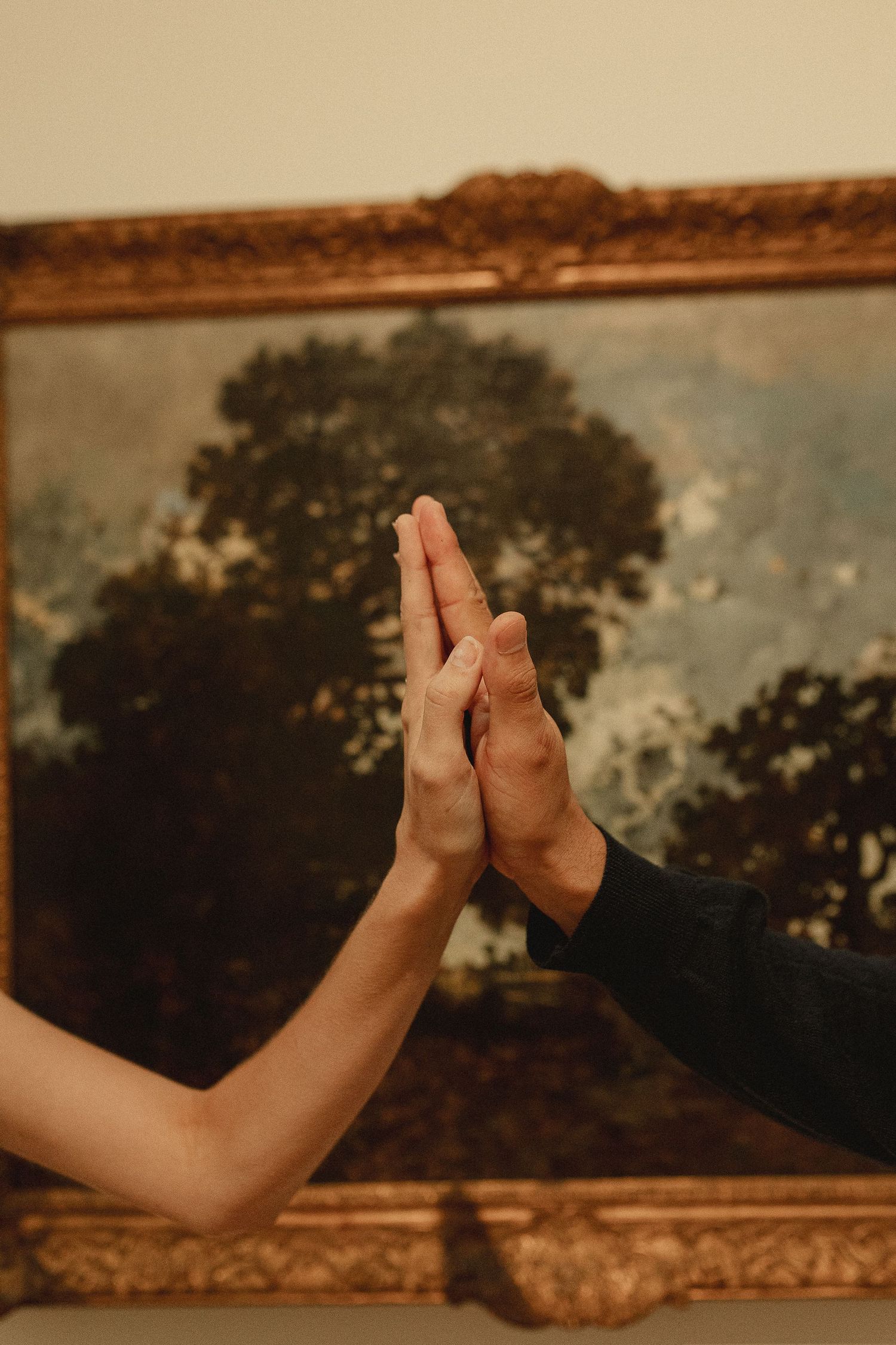 Two hands touching palm-to-palm in front of a classical landscape painting in a gold ornate frame.