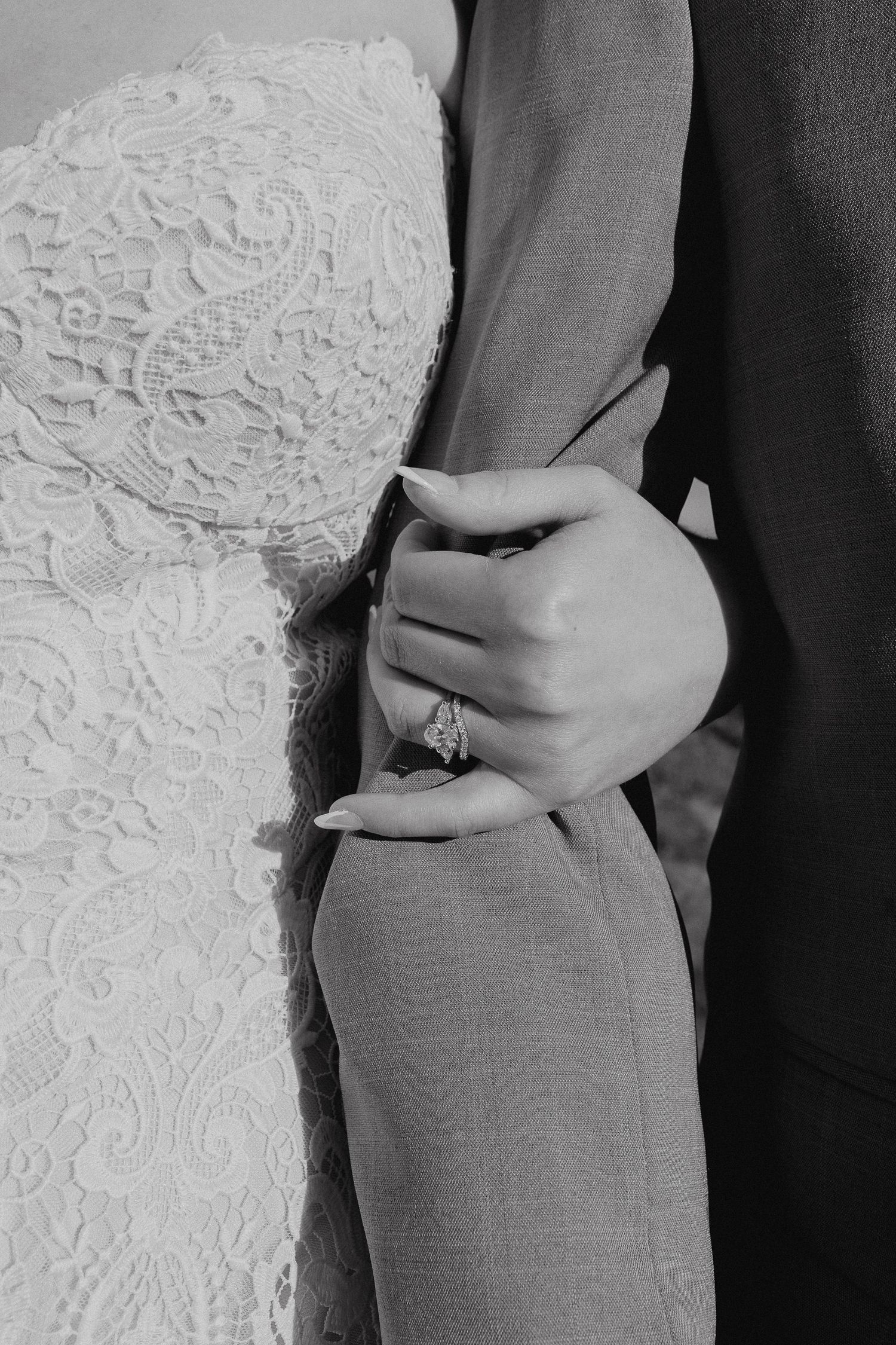 Black and white photo of bride in lace gown holding groom's arm, showcasing elegant diamond engagement ring.