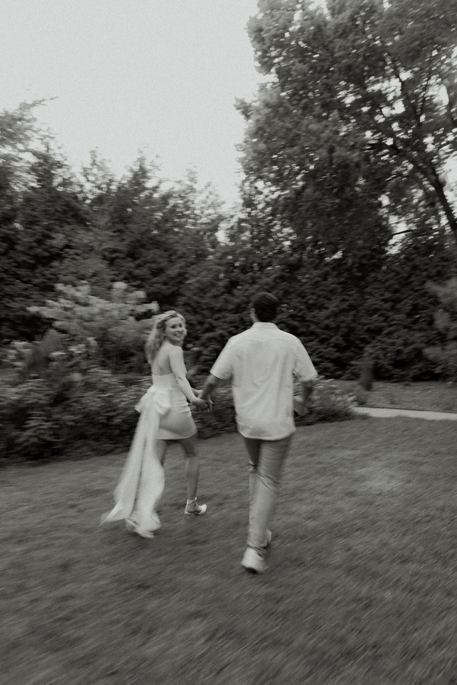 Black and white photo of a bride and groom holding hands, running through a lush garden on their wedding day.