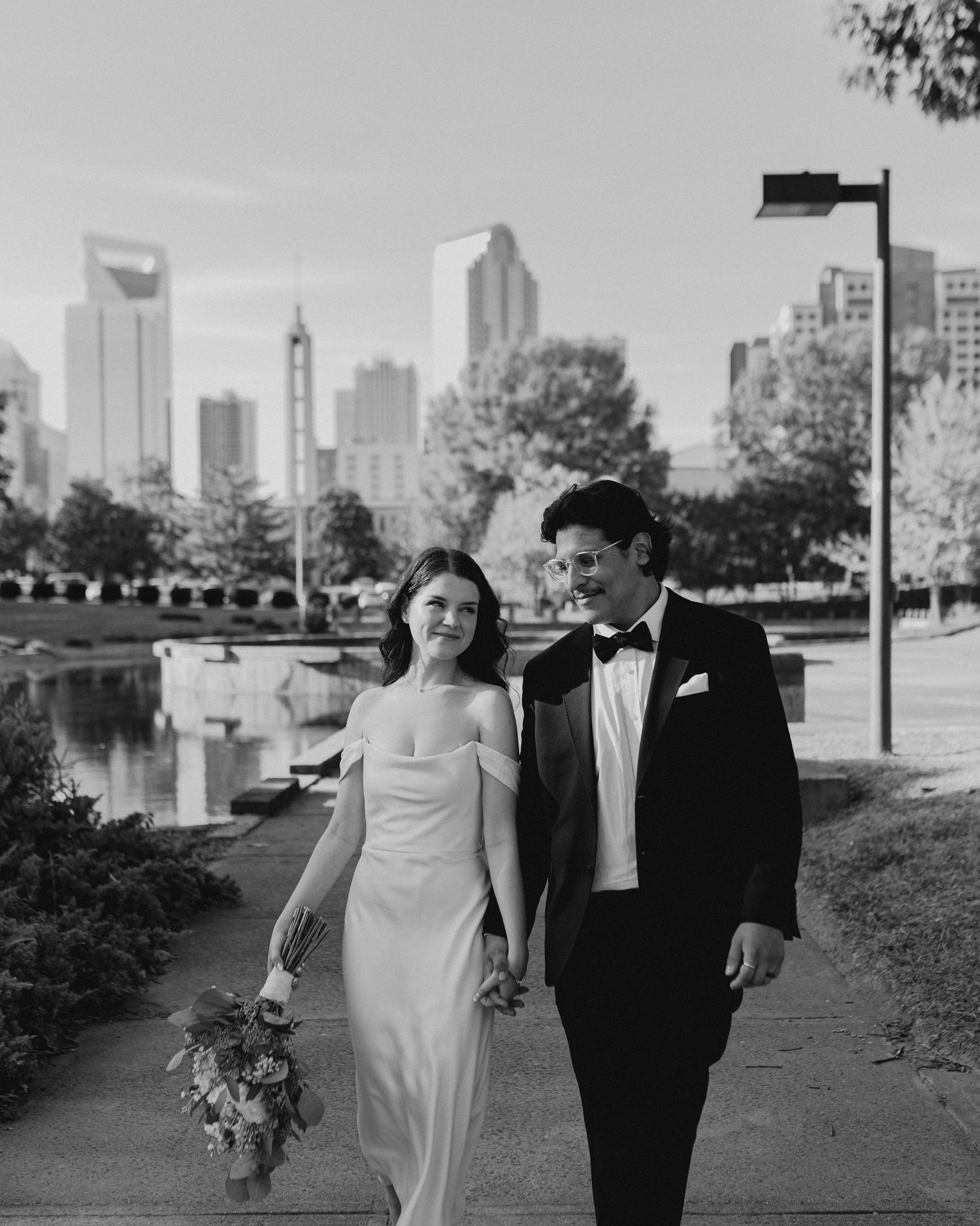 A couple walks along a city path with modern skyscrapers and water features in the background.