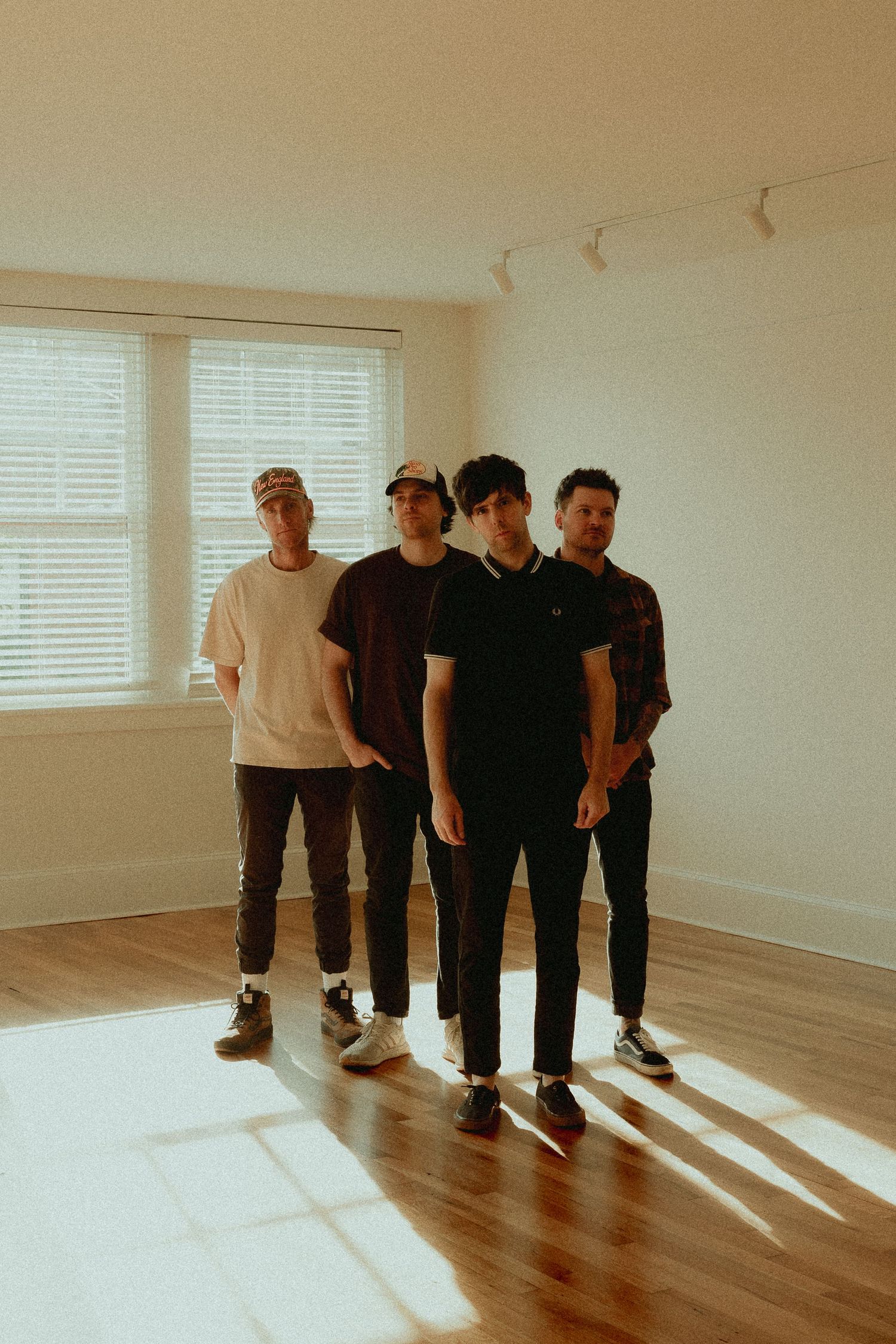 Four young men standing in a sunlit empty room with hardwood floors and white walls.