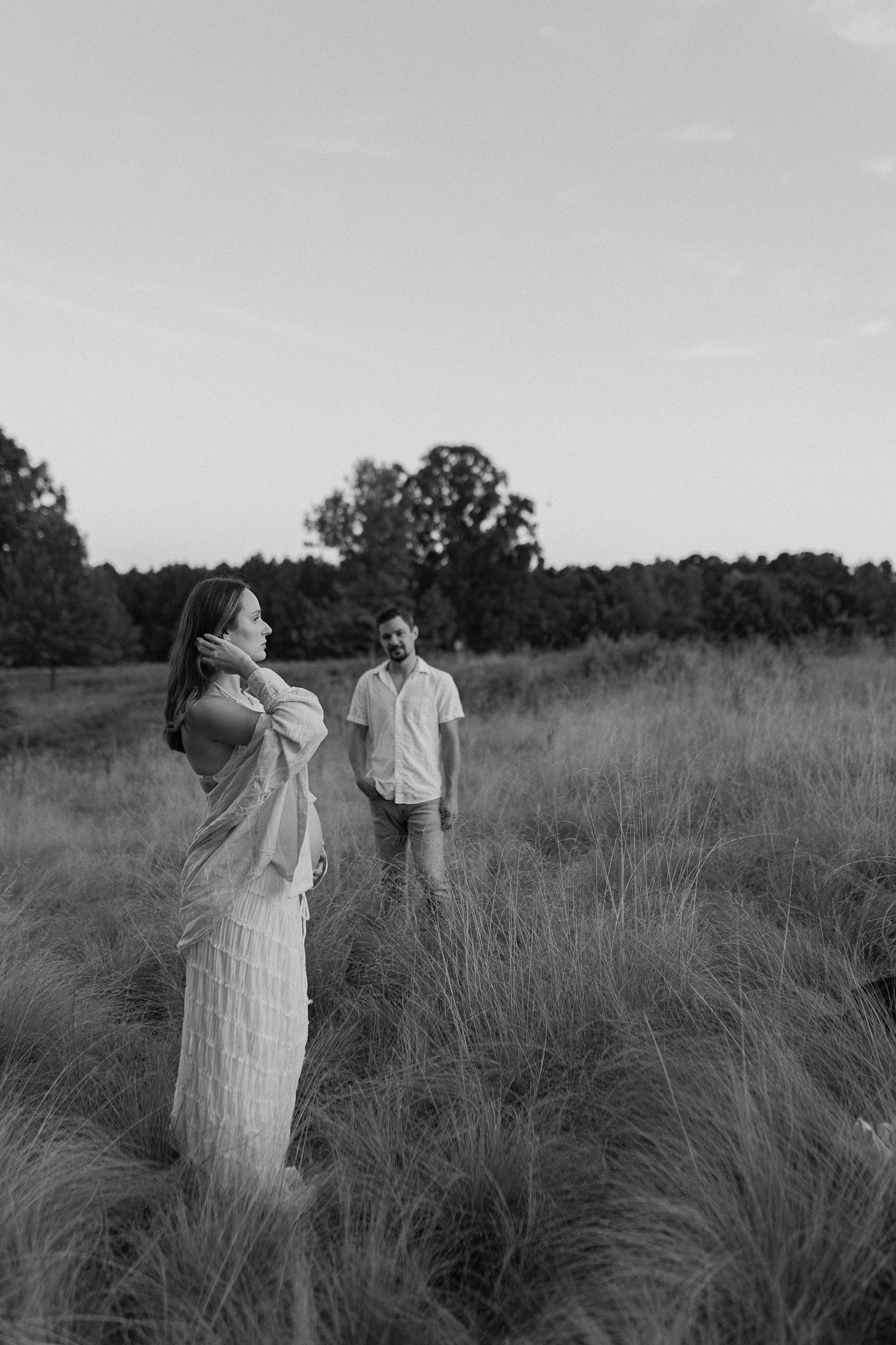 Black and white maternity portrait in tall grass field with trees in background.