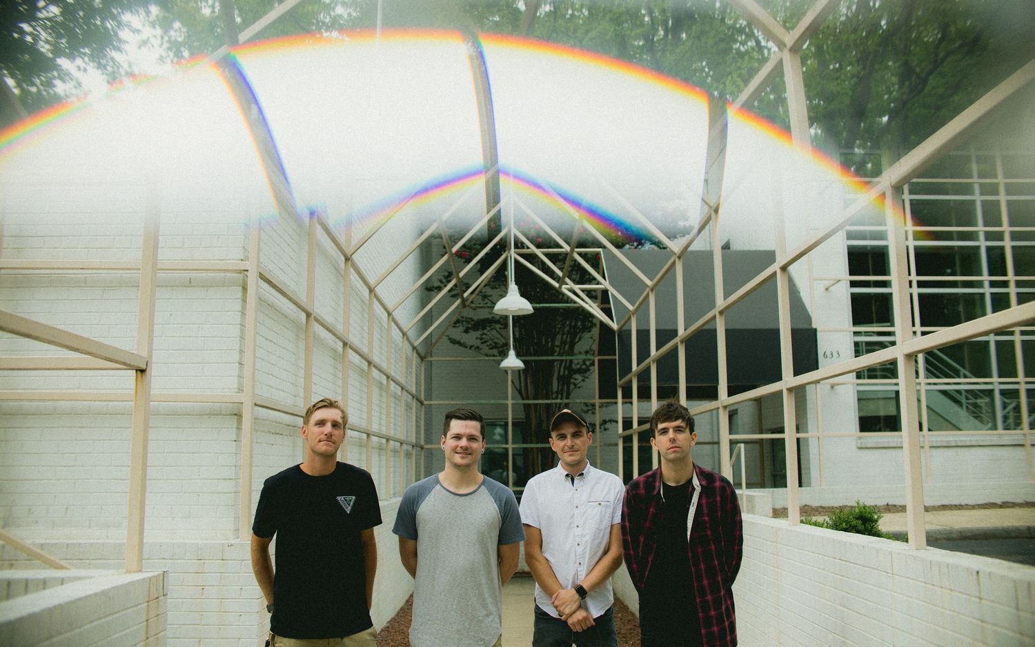 Four band members stand under a greenhouse-style structure with rainbow light effects.
