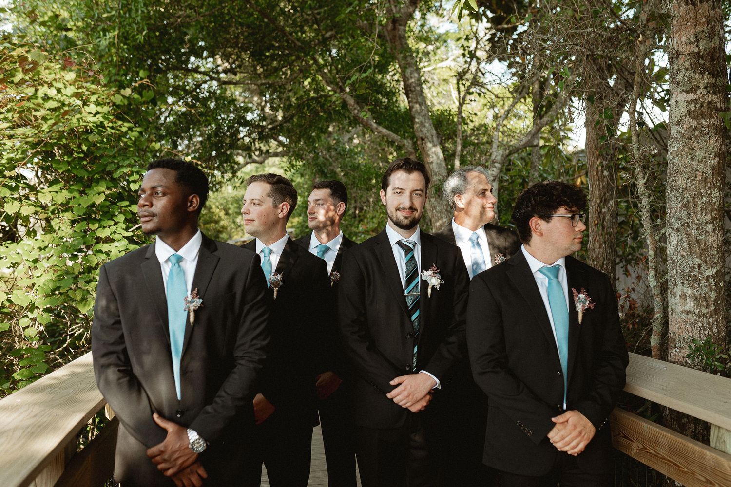 Wedding party in black suits with turquoise ties posed outdoors among trees.